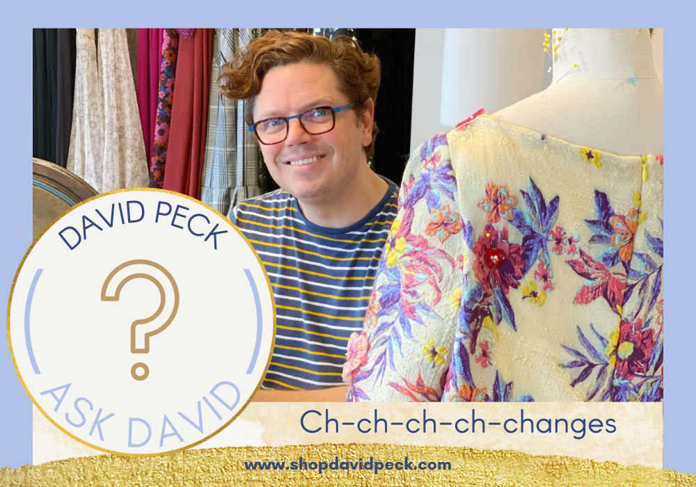 David smiling at the camera while sewing a dress. Man with curly hair in glasses and a striped shirt.