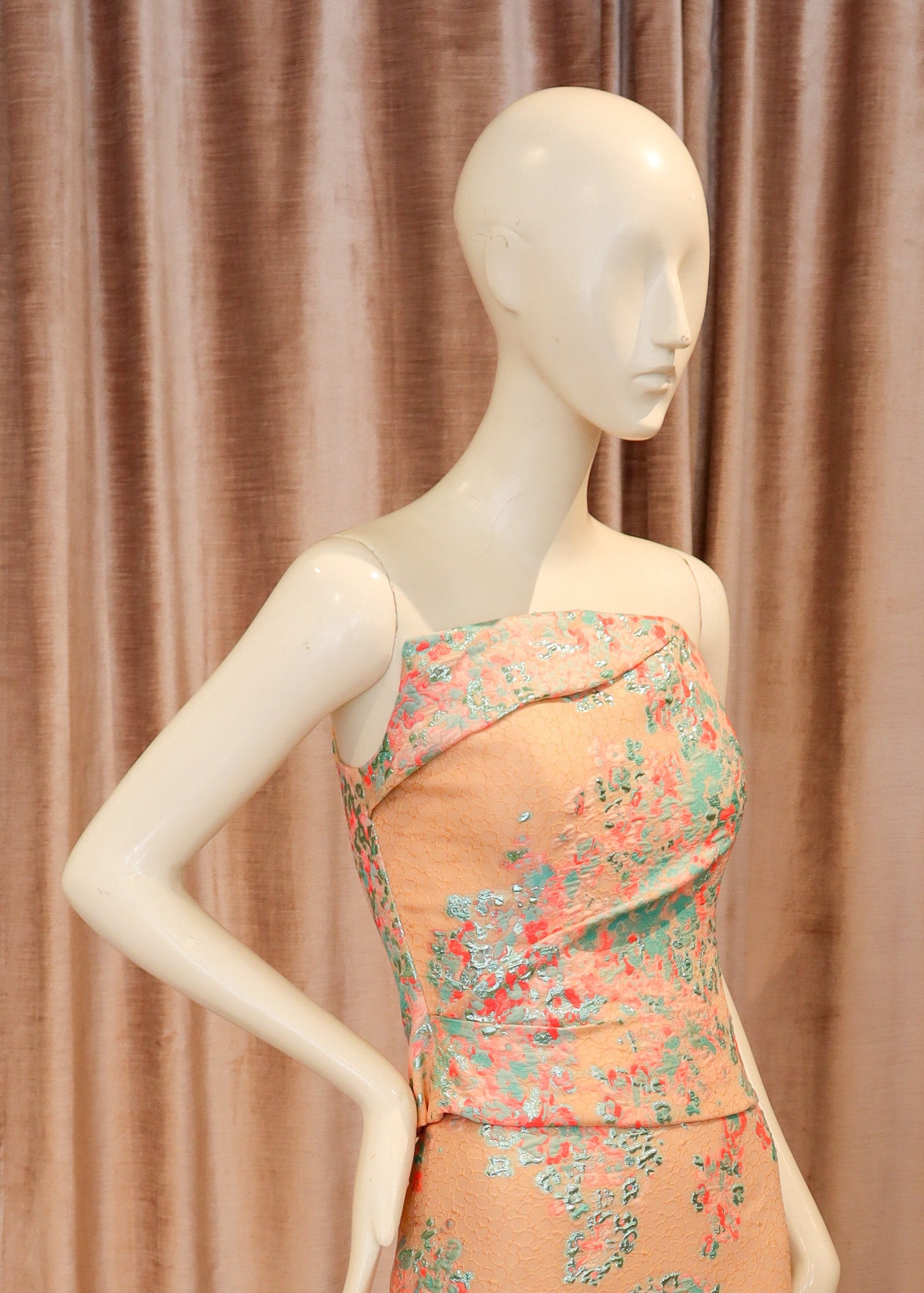 A peach-colored strapless evening gown with a pink and teal floral pattern, displayed on a white mannequin against a dusty pink velvet curtain and wooden parquet floor.