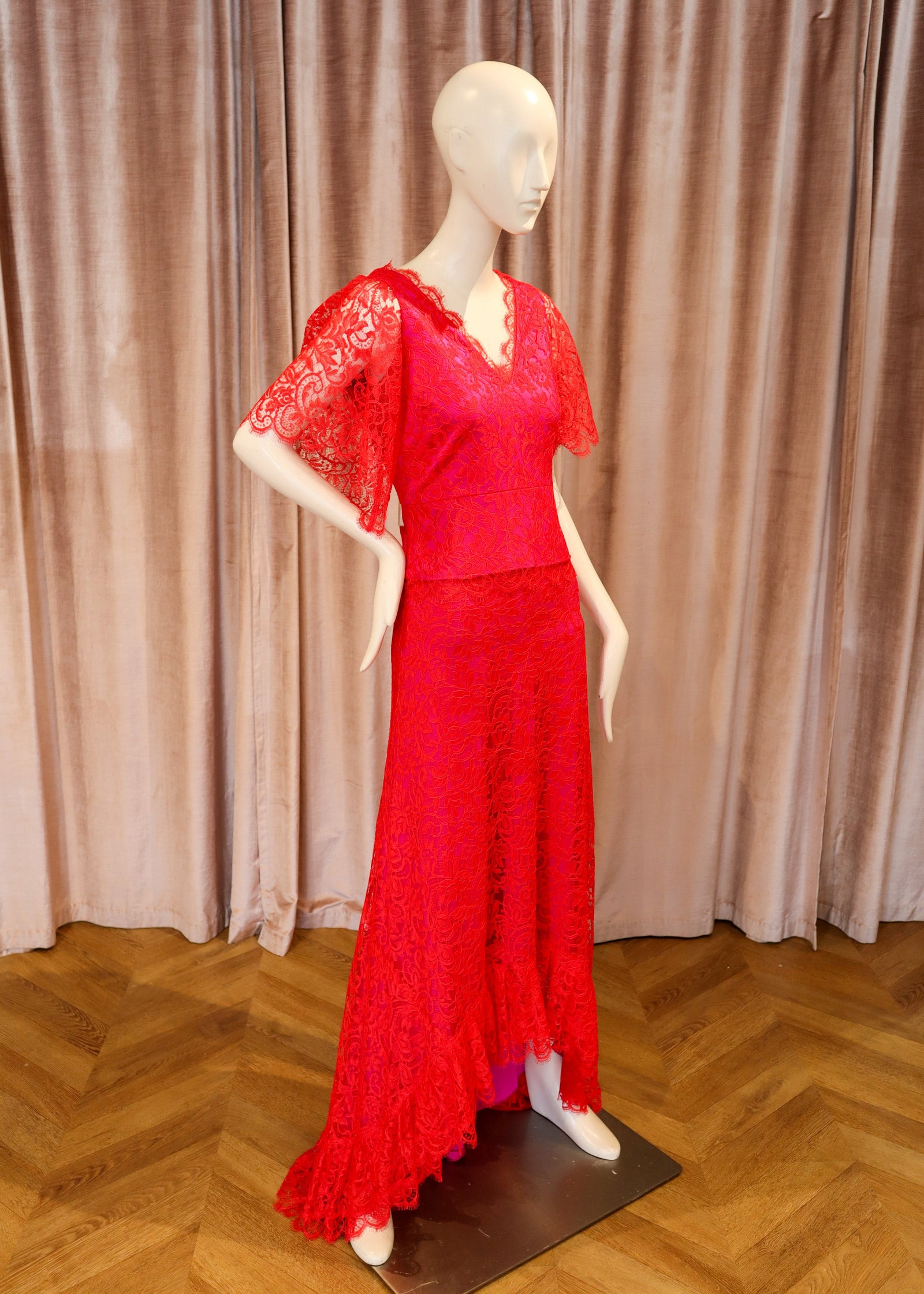 A bright red lace dress displayed on a white mannequin, set against a backdrop of draped mauve curtains and a wooden herringbone floor.