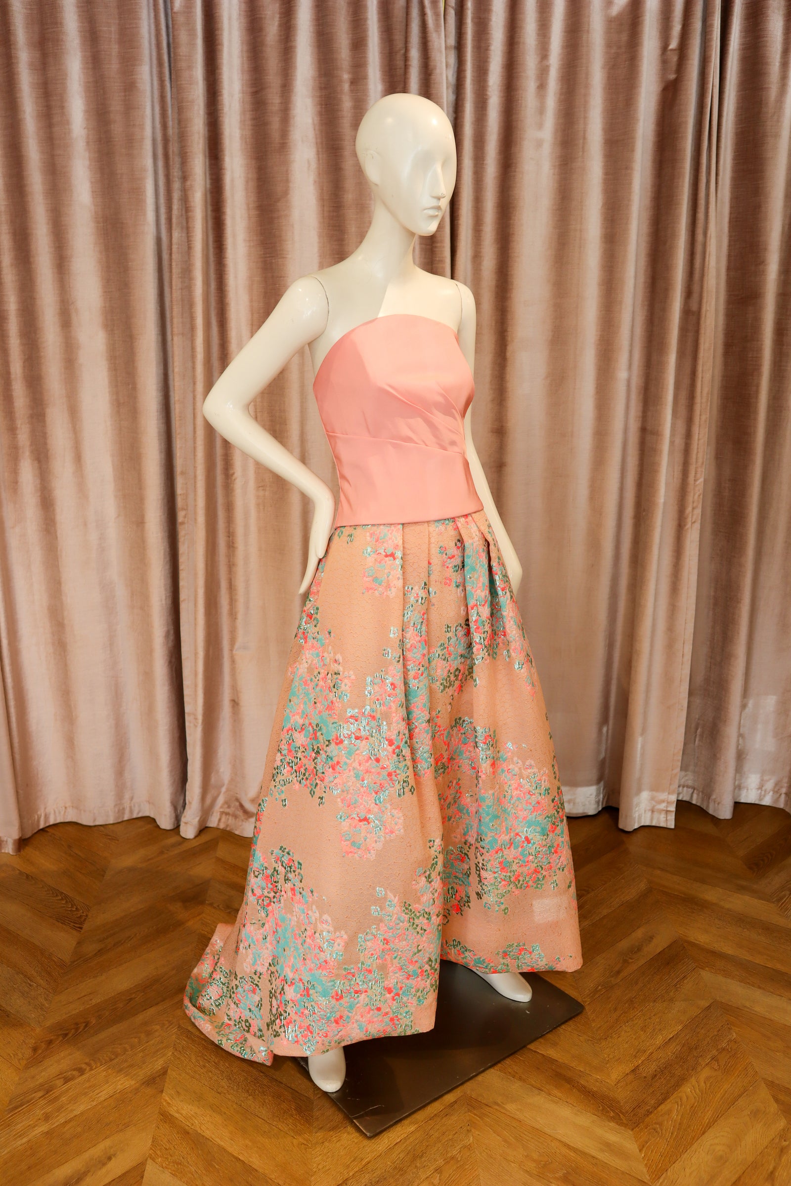 A peach-colored strapless bustier top displayed on a cream mannequin, paired with a beige satin skirt, set against a brown curtain backdrop.