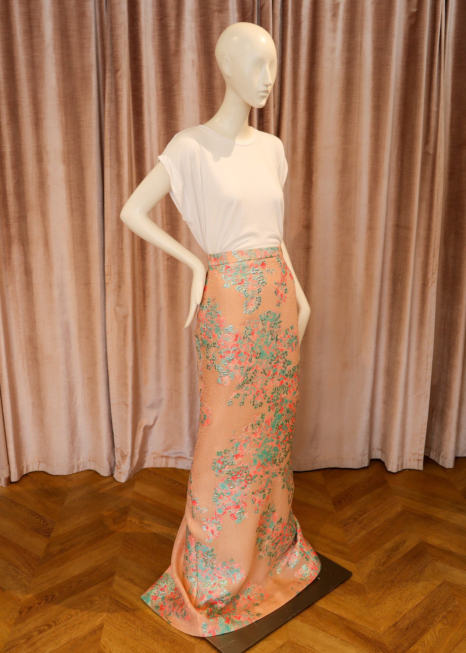 A mannequin wearing a white short-sleeve top and a peach maxi skirt with a green and pink floral print, set against a pink velvet curtain and a wooden parquet floor.