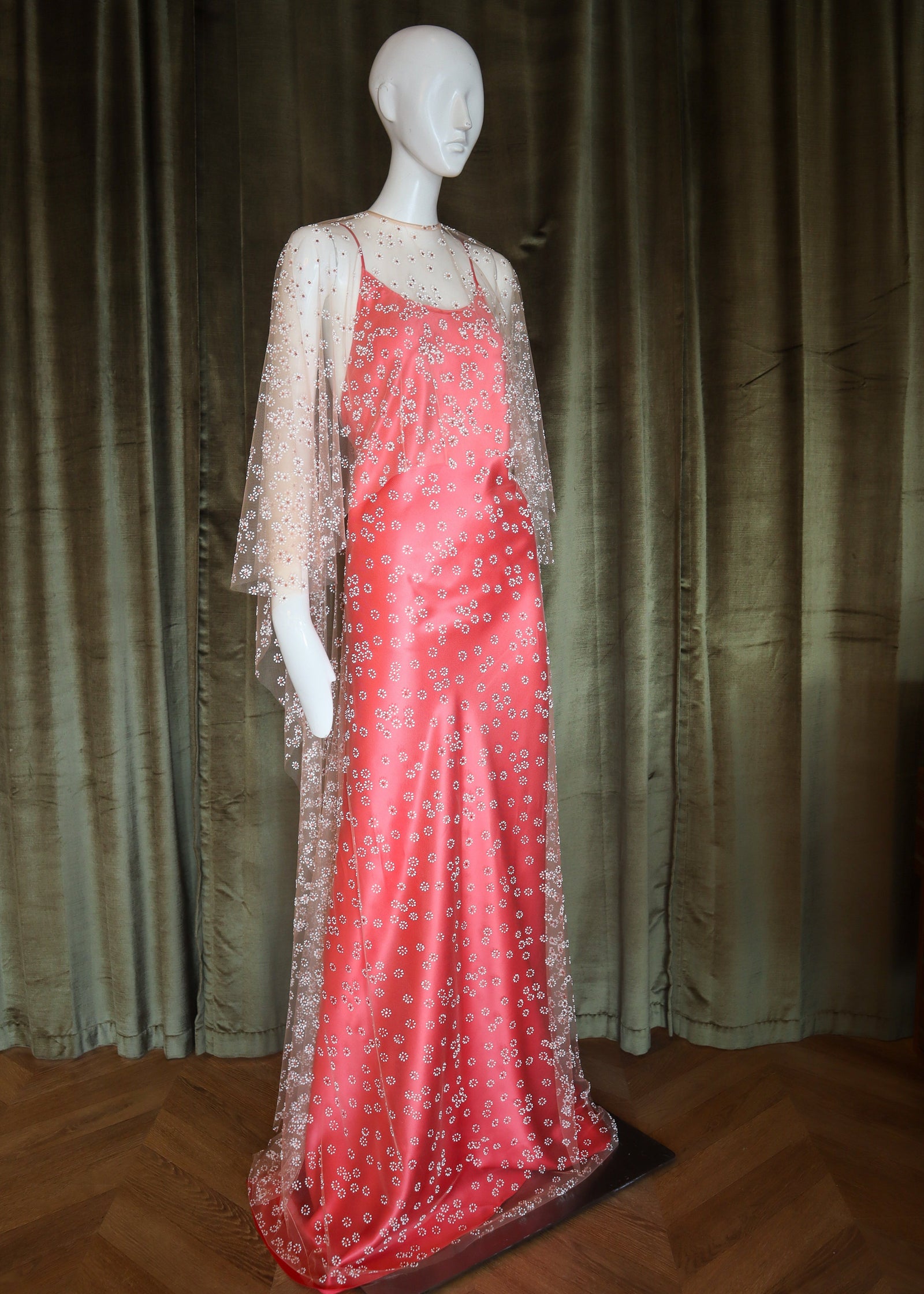 Mannequin wearing a red evening gown with a sheer overlay against a dark curtain background