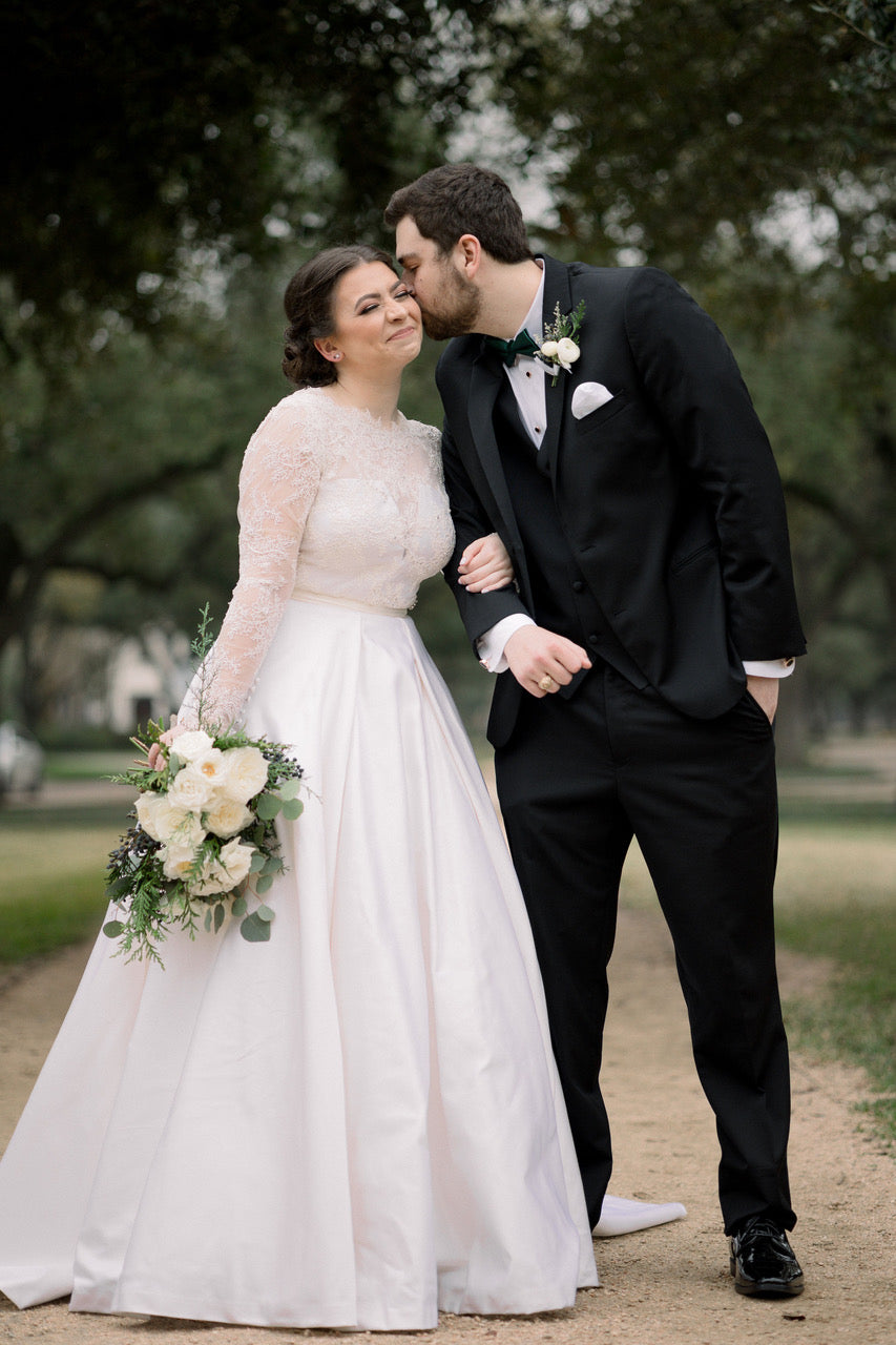 Kasey French Raley in her custom long sleeve lace wedding dress by David Peck receiving a kiss on the cheek by her husband at her outdoor wedding.