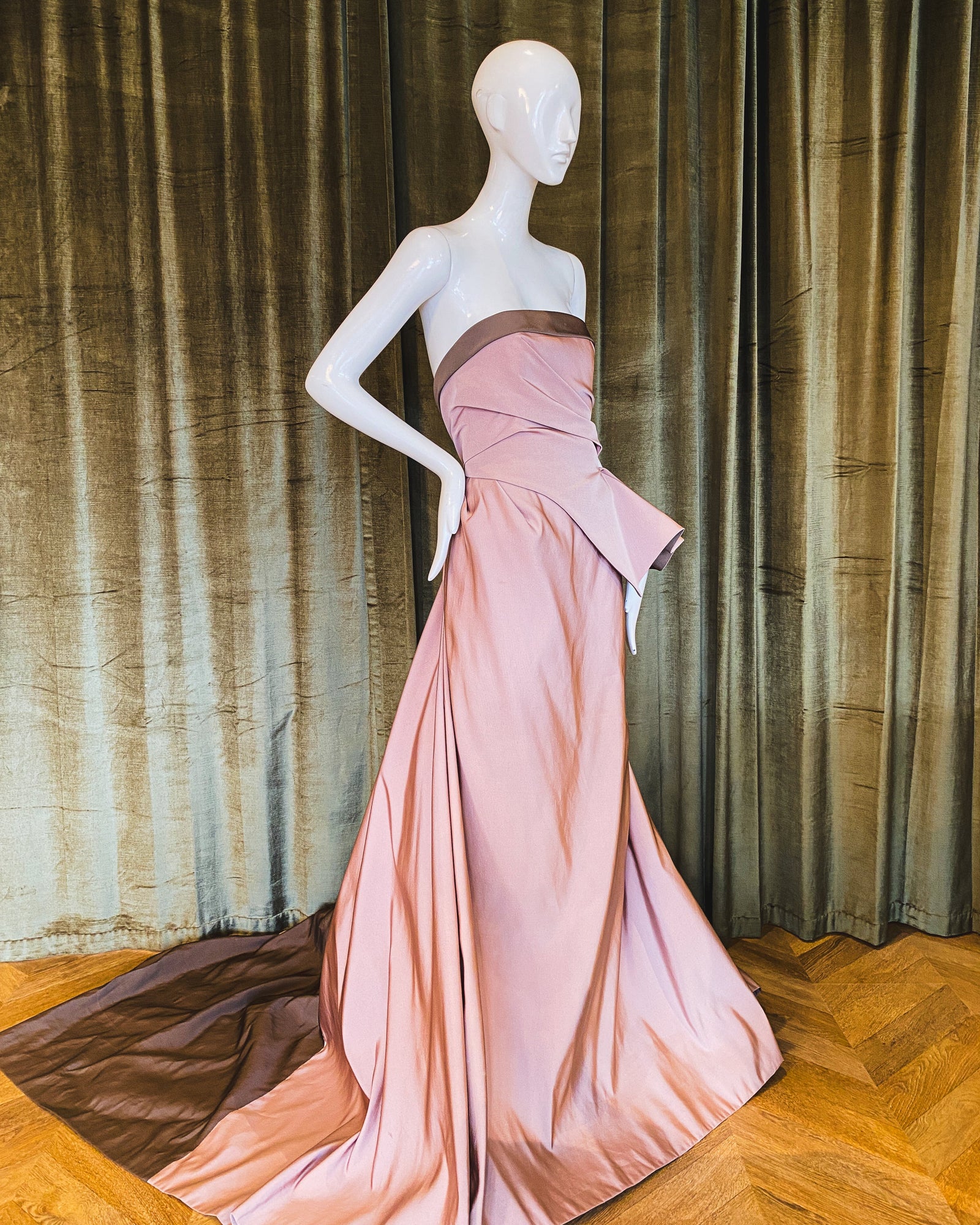 A mannequin displaying a pink silk faille gown with a boned bustier and a sweeping train set against a curtain backdrop. designed by David Peck