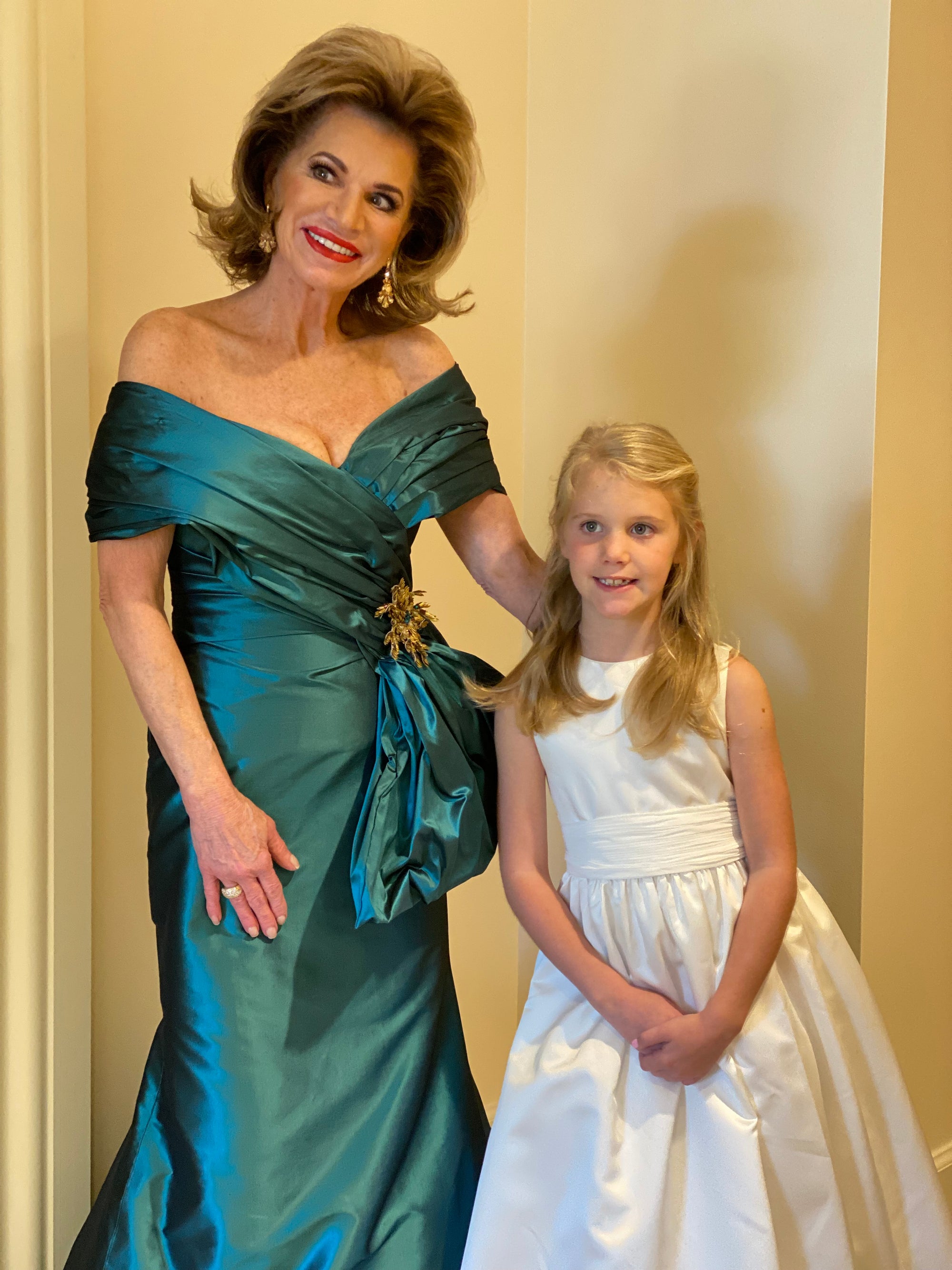 Debra Laws in her custom emerald green, off the shoulder, and wrap mother of the bride dress by David Peck smiling and posing next to a flower girl.