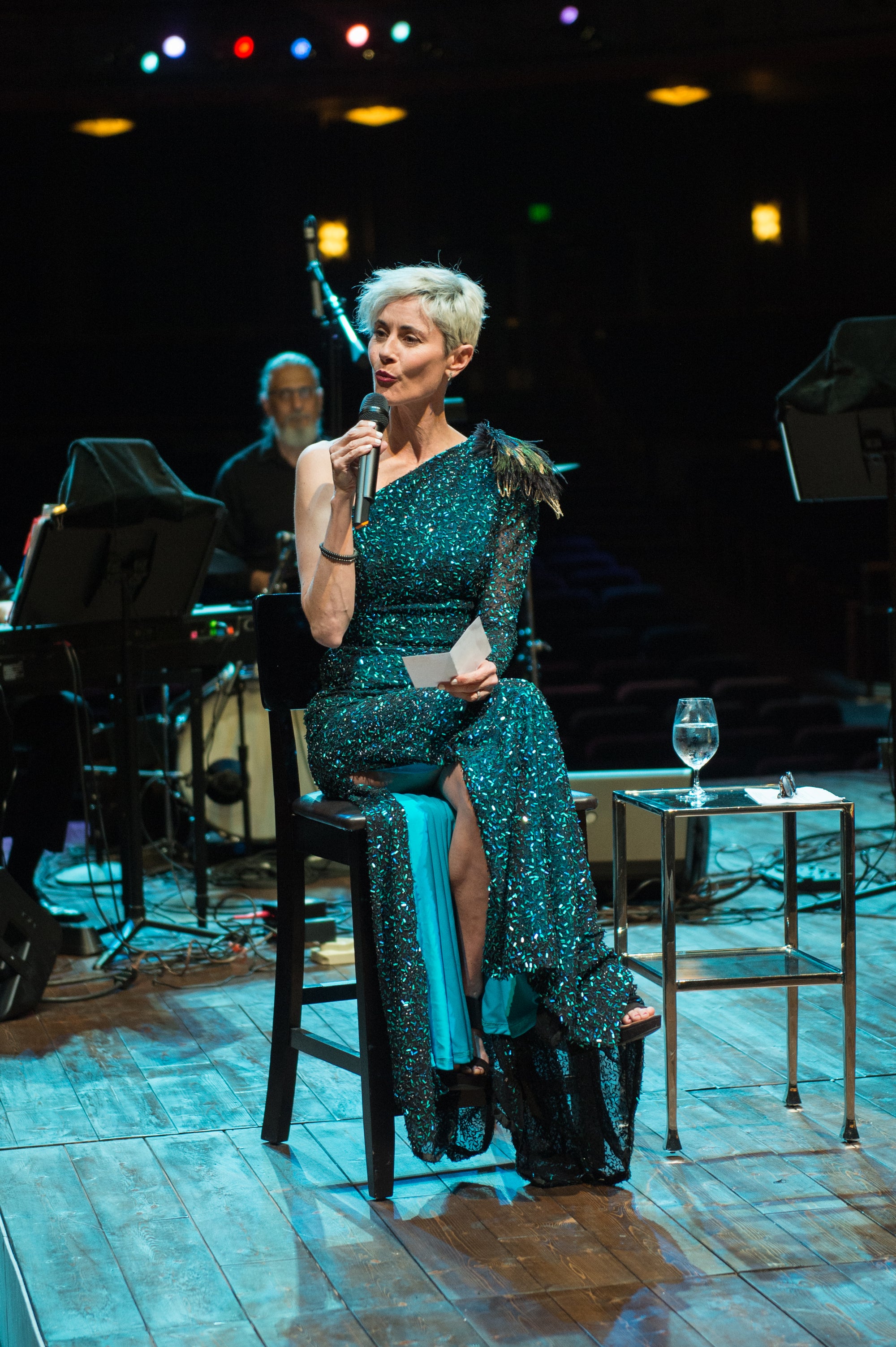 Beth Malone, a Tony Award Nominee, wearing a custom one shoulder and sequin gown by David Peck sitting down and singing at a venue.