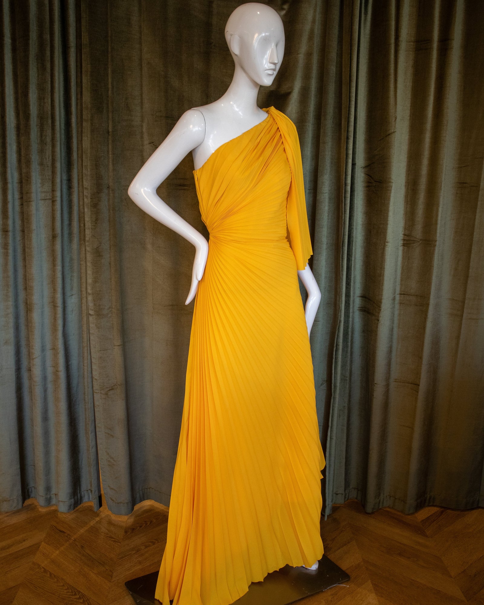A marigold yellow gown with pleated detail displayed on a mannequin against a neutral backdrop.
