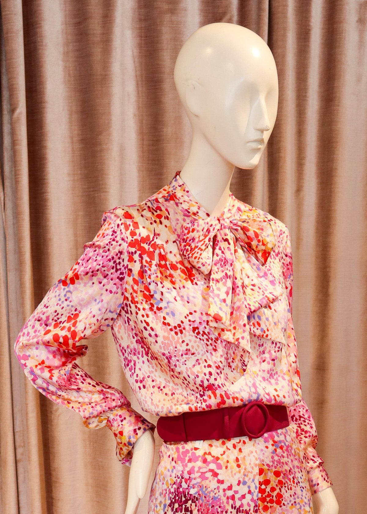 A mannequin wearing a matching floral long-sleeve blouse and wide-leg trousers set in pink, red, orange, and white, with a dark red belt, standing against a pink velvet curtain and wooden floor.