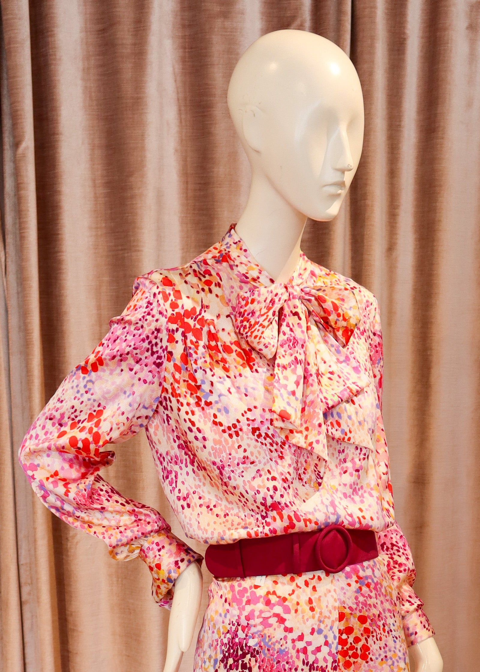 A mannequin wearing a matching floral long-sleeve blouse and wide-leg trousers set in pink, red, orange, and white, with a dark red belt, standing against a pink velvet curtain and wooden floor.