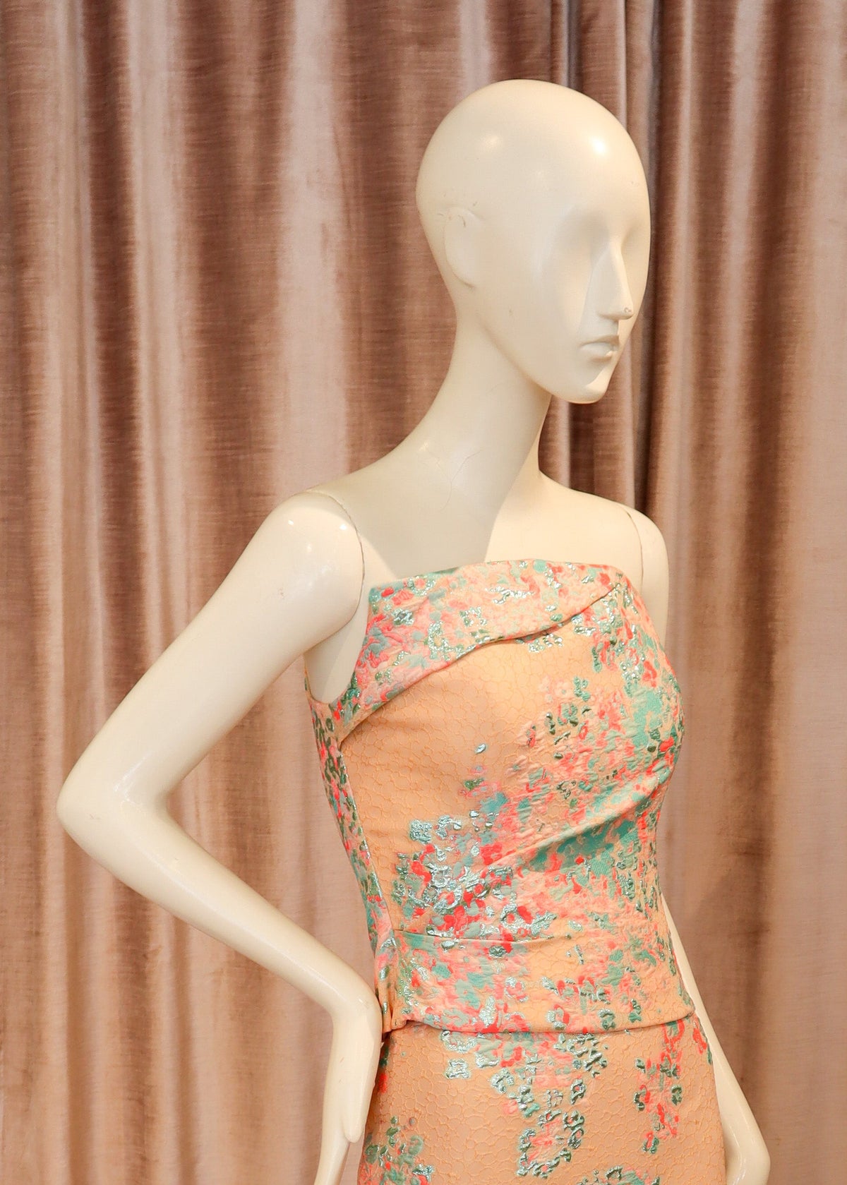 A peach-colored strapless evening gown with a pink and teal floral pattern, displayed on a white mannequin against a dusty pink velvet curtain and wooden parquet floor.