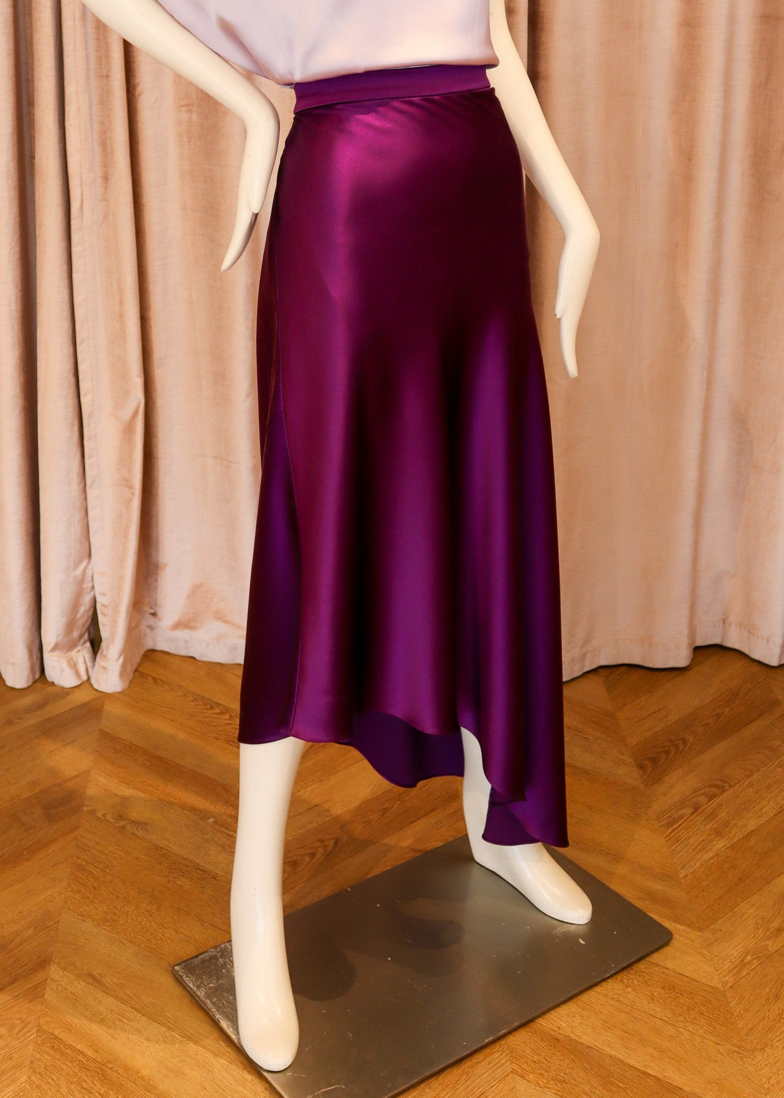 A mannequin wearing a two-piece outfit consisting of a light lavender off-the-shoulder top and a shiny dark purple satin midi skirt, displayed against a backdrop of pinkish-beige velvet curtains and a wooden herringbone floor.
