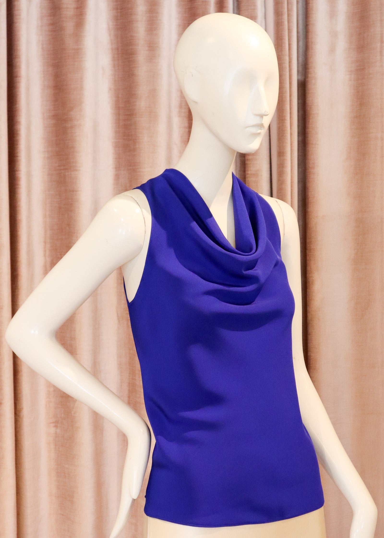 A sleeveless cowl-neck top in vivid royal blue displayed on a cream mannequin, set against a backdrop of draped pinkish-beige curtains.