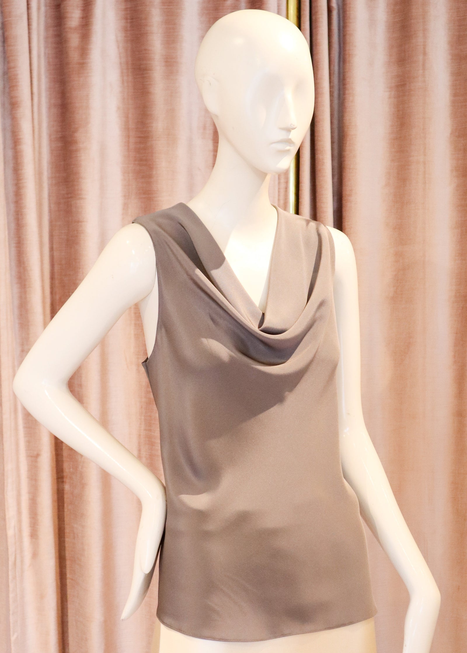 A sleeveless cowl-neck top in taupe brown displayed on a cream mannequin against a draped mauve-pink curtain background.