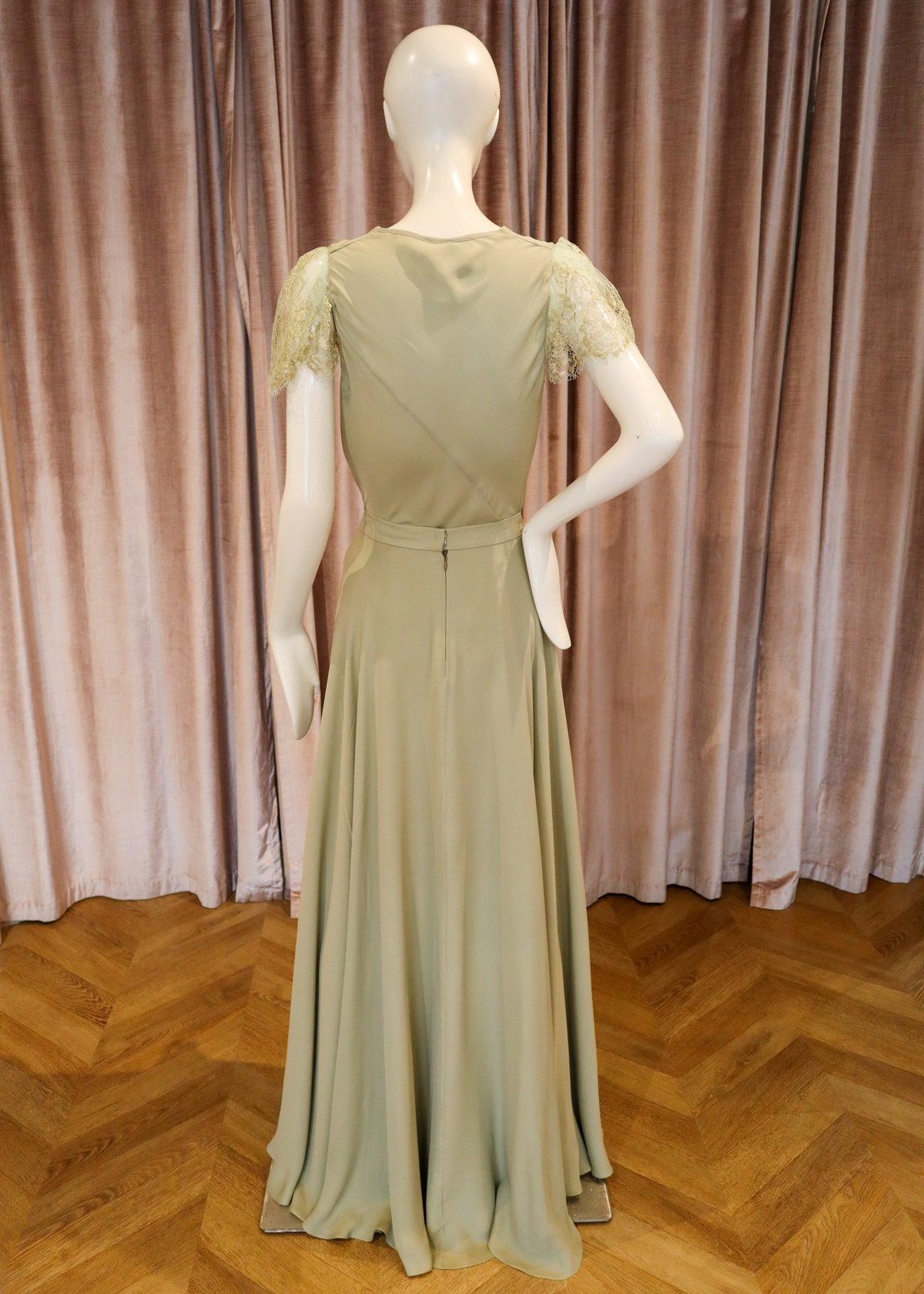 A floor-length sage green evening gown displayed on a white mannequin from the back, featuring lace short sleeves and a wrap-style back, set against a pink velvet curtain and wooden parquet floor.