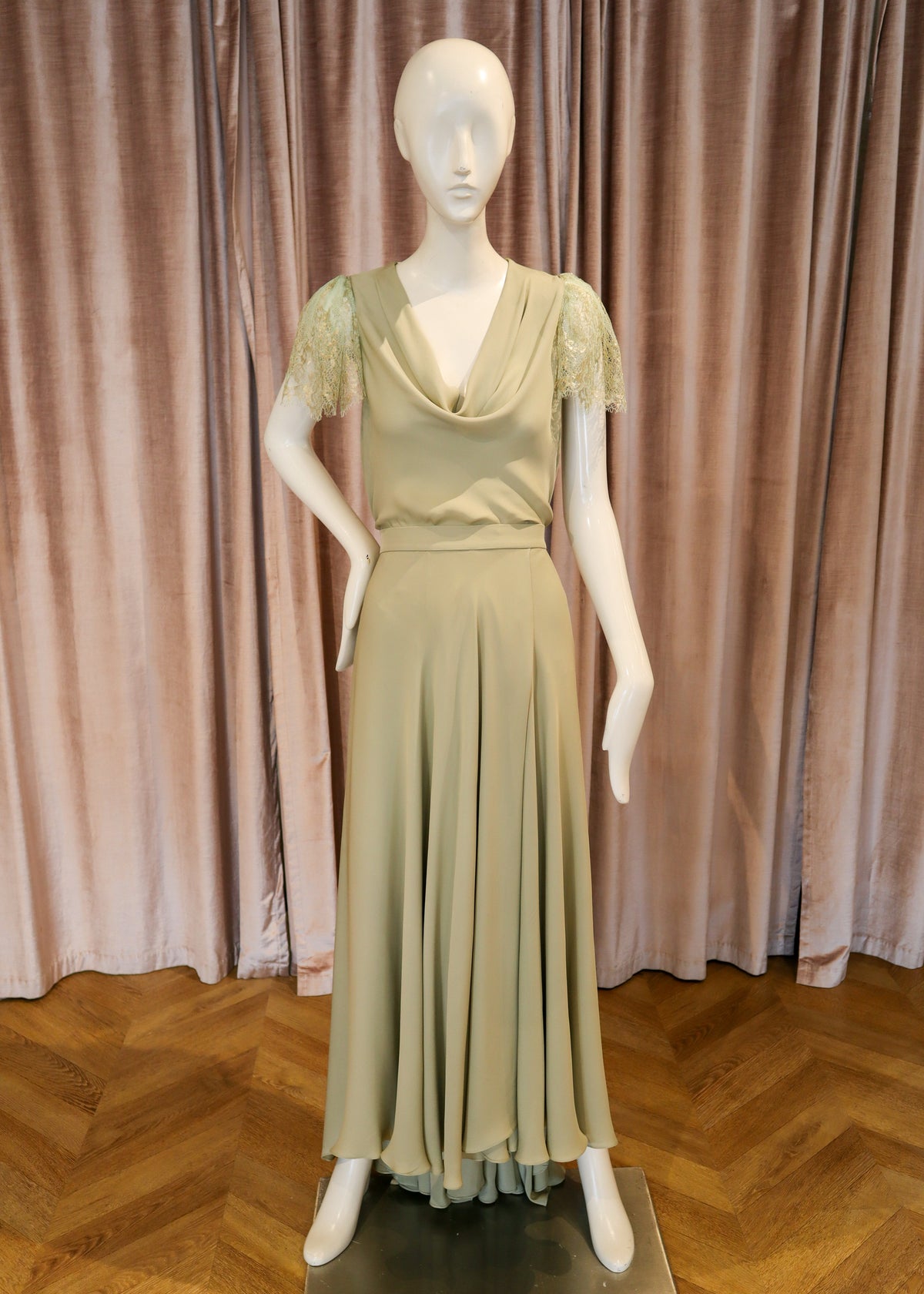 A cream-colored maxi dress with short lace sleeves and a cowl neckline, displayed on a white mannequin against a pink velvet curtain and wooden parquet floor.