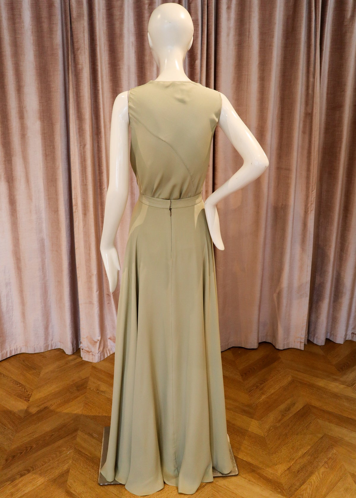 A sleeveless floor-length dress displayed on a white mannequin, shown from the back against a pink velvet curtain and wooden herringbone floor.