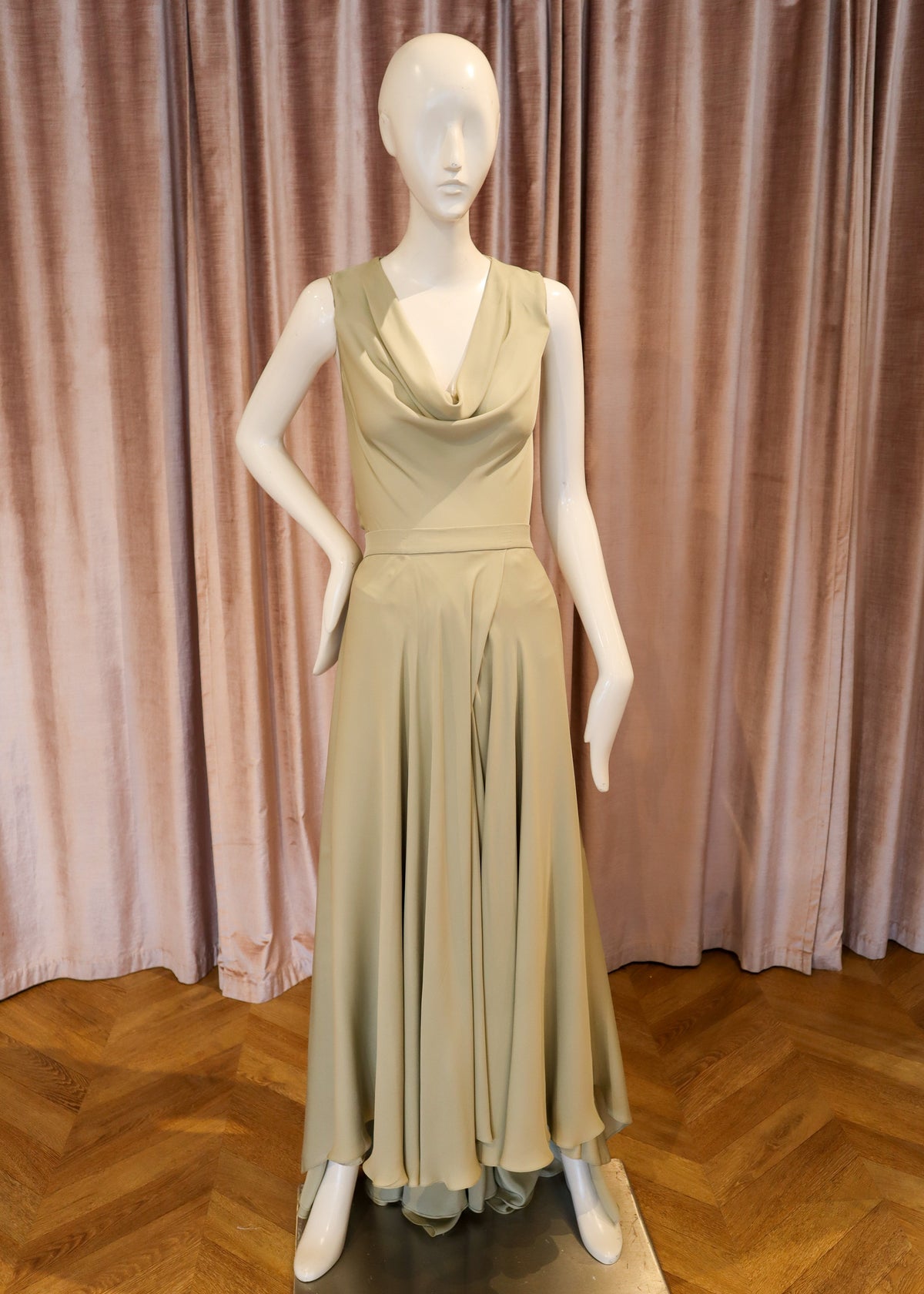 A sleeveless cowl-neck maxi dress in a light beige or sage green shade, displayed on a white mannequin standing on a metallic platform in front of pink curtains and a wooden herringbone floor.