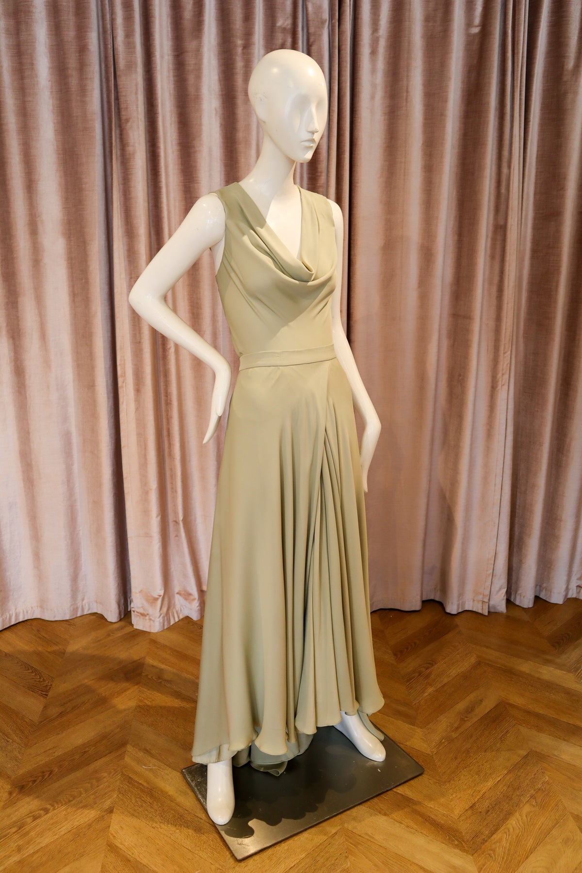 A sleeveless, cowl-neck, floor-length dress in a light sage green or khaki shade, displayed on a white mannequin. The dress features a draped neckline, a defined waist, and a flowing skirt.