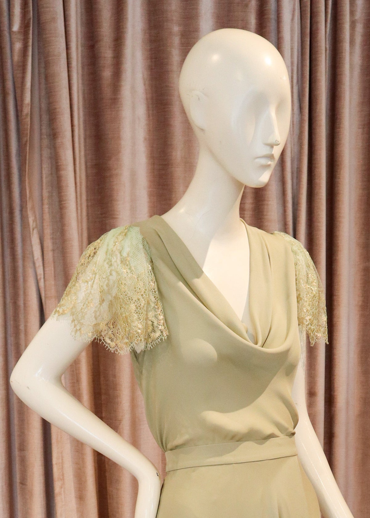 A floor-length sage green dress displayed on a white mannequin, featuring short sheer lace sleeves and a draped neckline, set against a pink velvet curtain and wooden parquet floor.