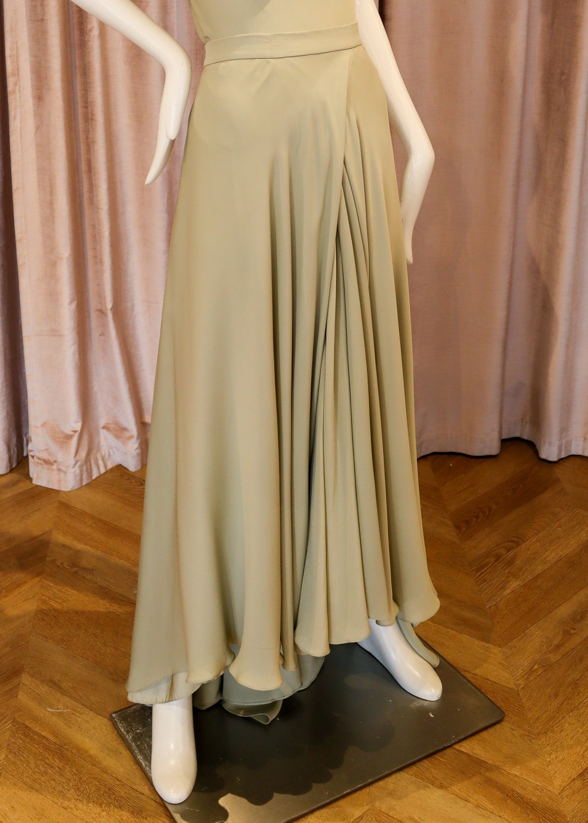 A sleeveless, cowl-neck, floor-length dress in a light sage green or khaki shade, displayed on a white mannequin. The dress features a draped neckline, a defined waist, and a flowing skirt.