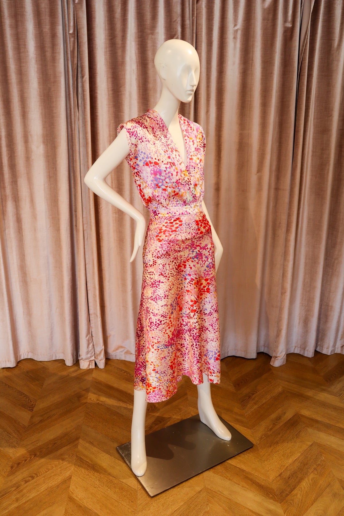 A sleeveless V-neck midi dress with a colorful abstract floral print, displayed on a white mannequin against a pink velvet curtain and wooden parquet floor.