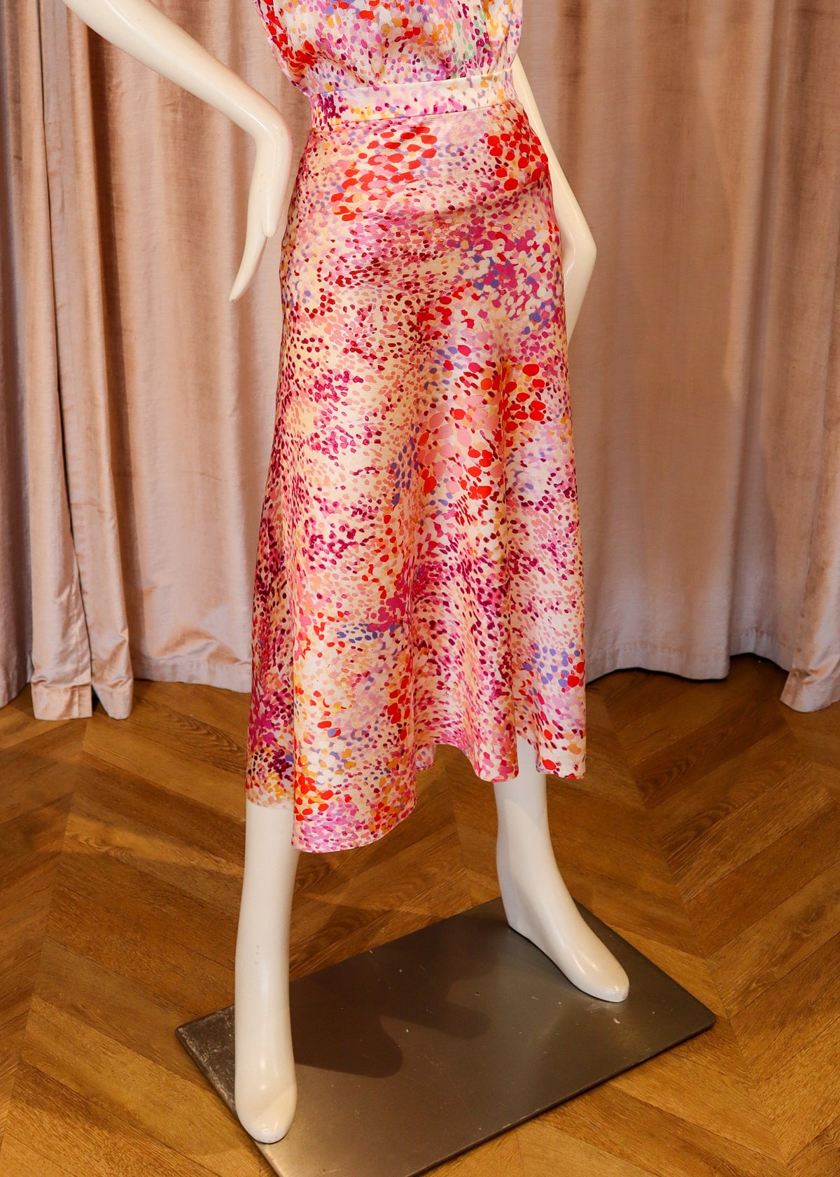 A sleeveless V-neck midi dress with a colorful abstract floral print, displayed on a white mannequin against a pink velvet curtain and wooden parquet floor.