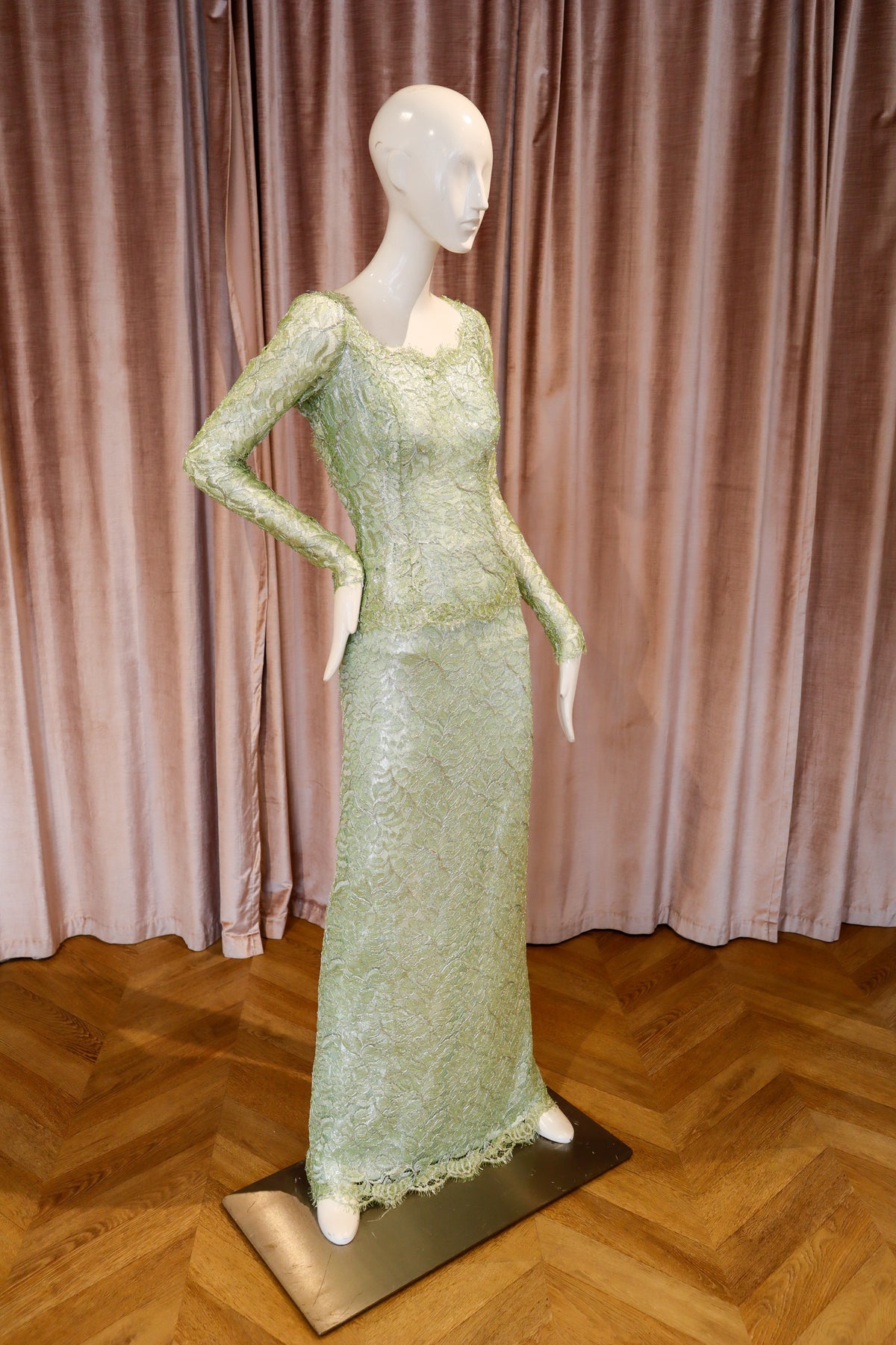 A floor-length light sage green lace gown displayed on a white mannequin, set against a backdrop of pinkish-beige velvet curtains and a wooden parquet floor.