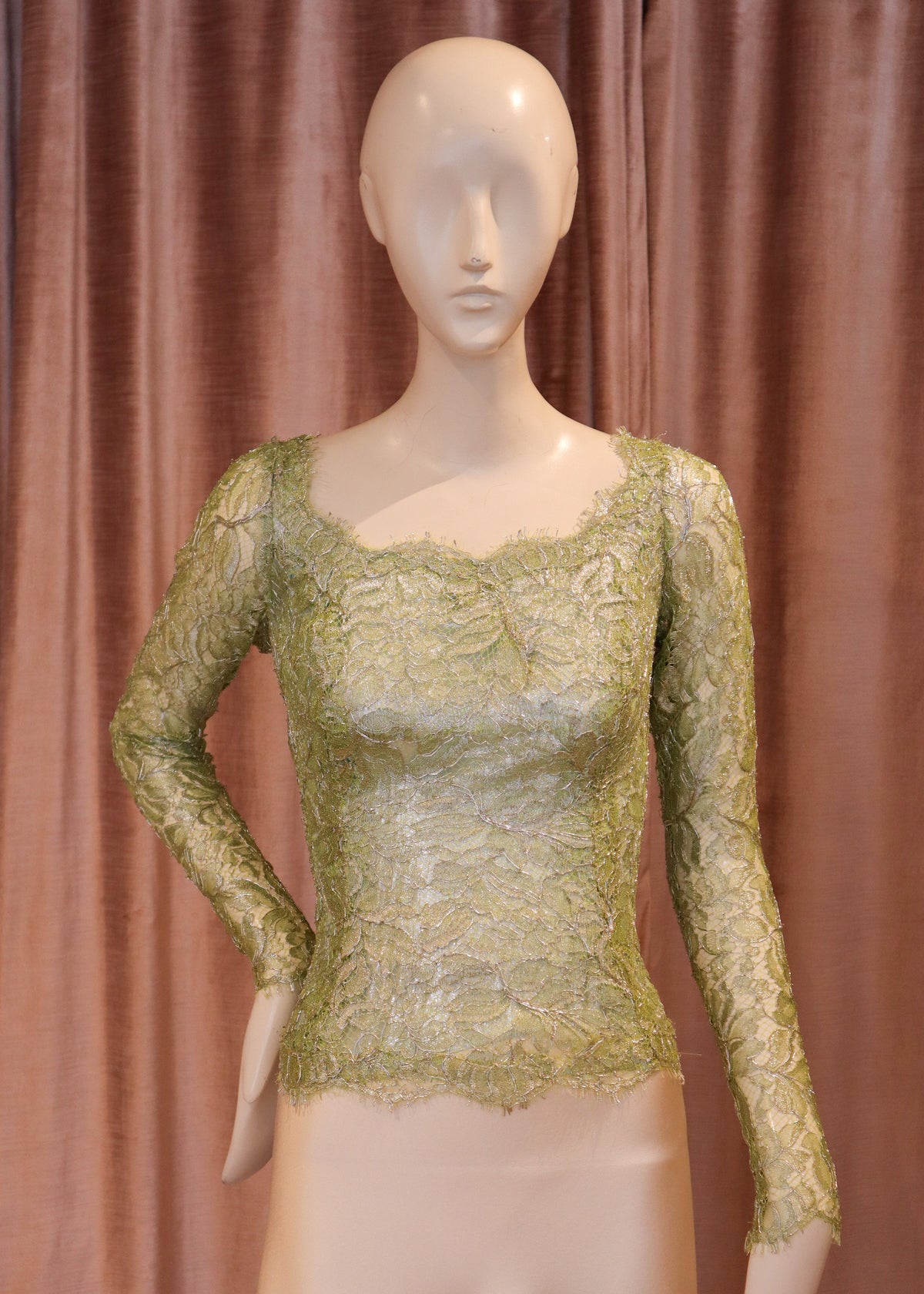 A long-sleeve lace top in a muted olive green color, displayed on a beige mannequin against a brown curtain backdrop.