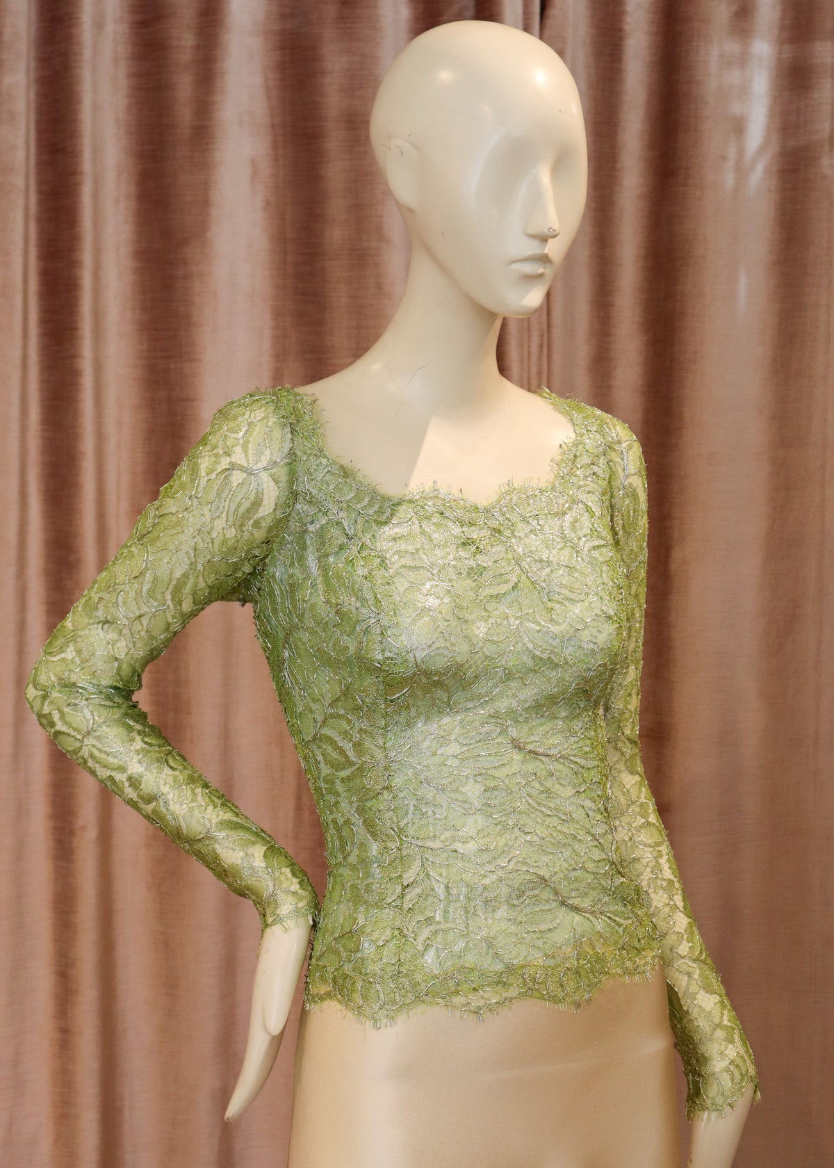 A long-sleeve lace top in light olive green displayed on a beige mannequin, set against a brown curtain backdrop.