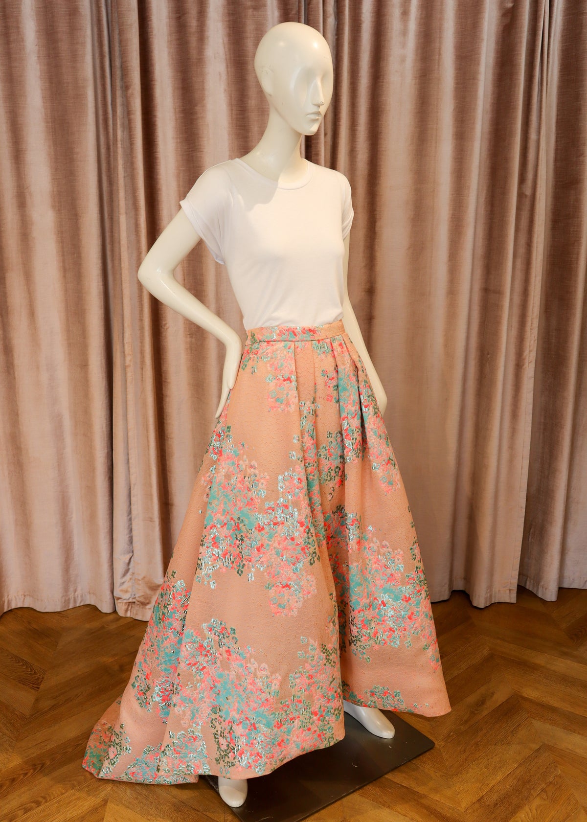 A white short-sleeve top paired with a high-waisted, full-length peach skirt featuring a colorful floral print, displayed on a white mannequin in front of pink curtains and a wooden floor.