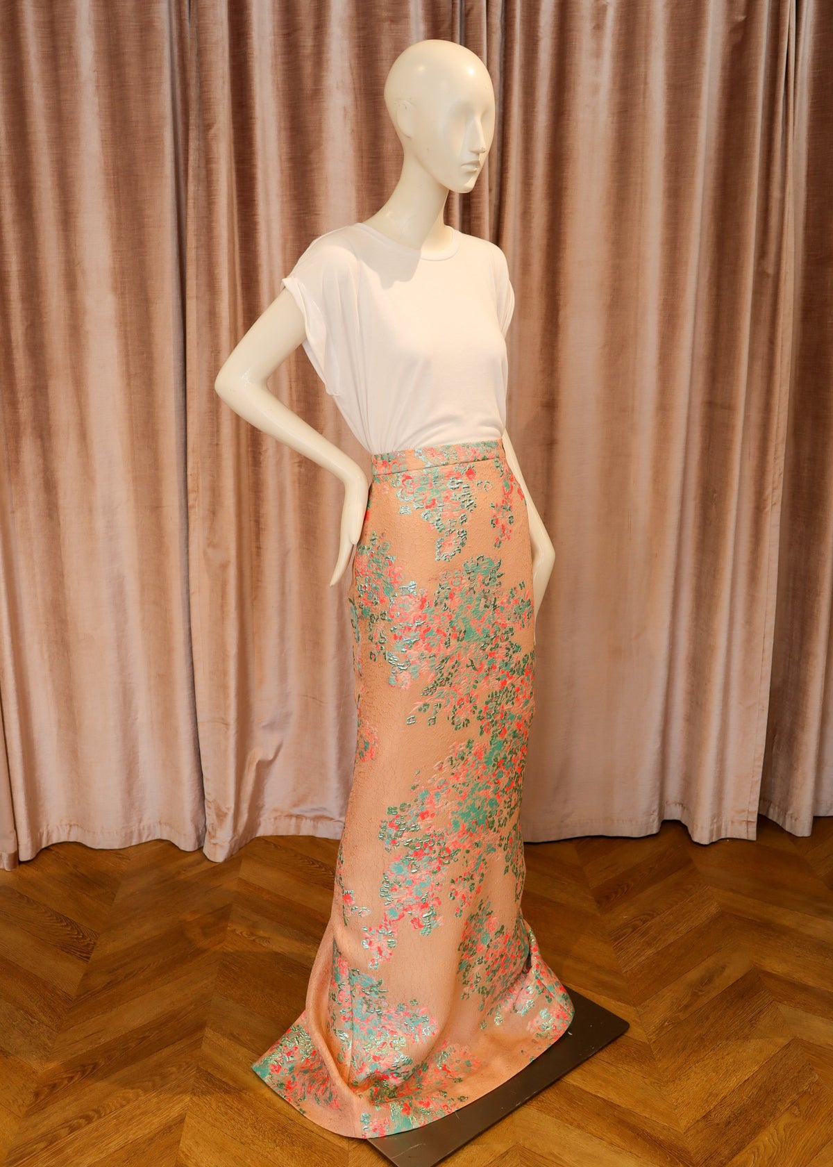 A mannequin wearing a white short-sleeve top and a peach maxi skirt with a green and pink floral print, set against a pink velvet curtain and a wooden parquet floor.