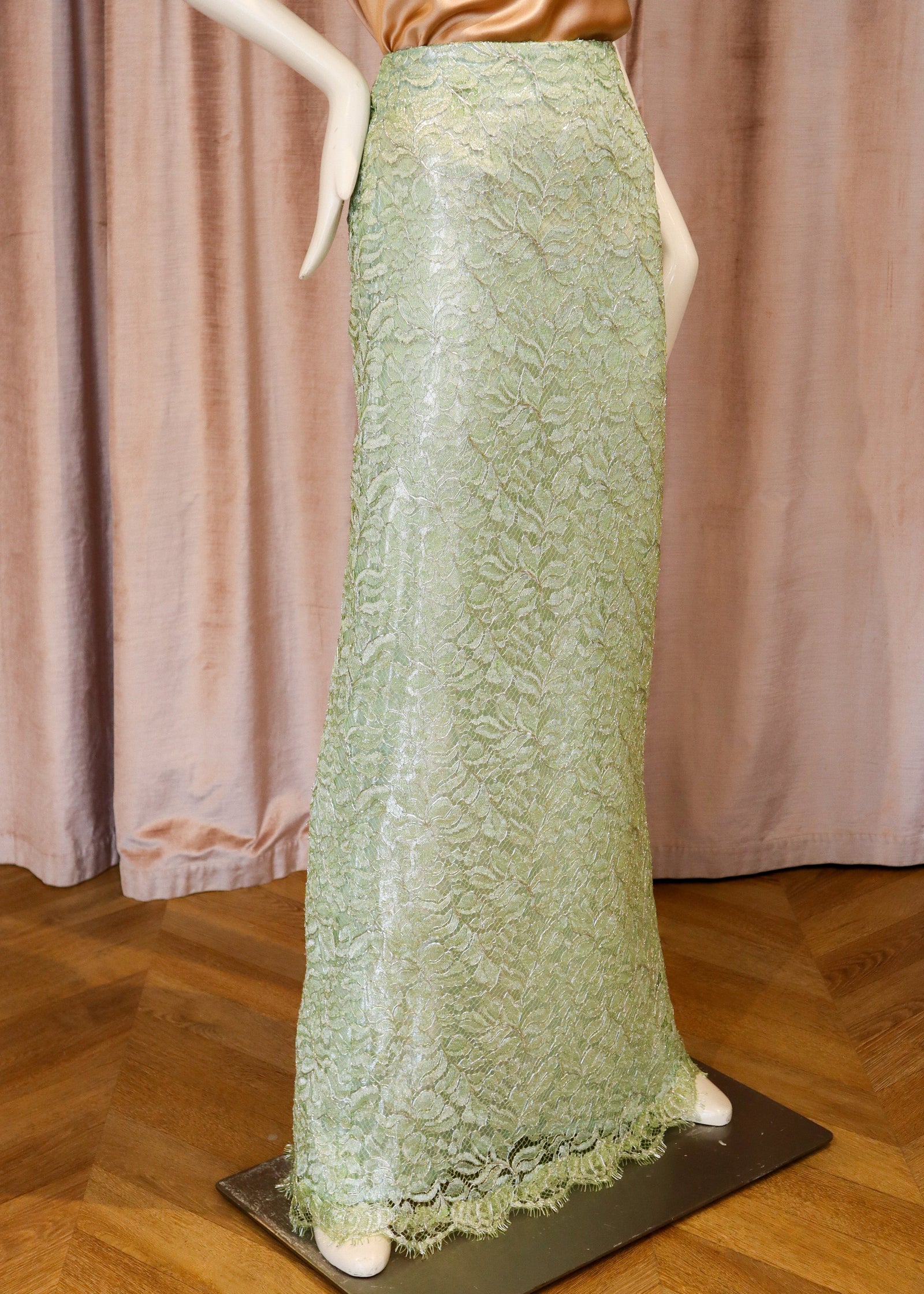 A mannequin wearing a long, floor-length light green lace skirt with a satin metallic top, set against a pink curtain backdrop and wooden floor.