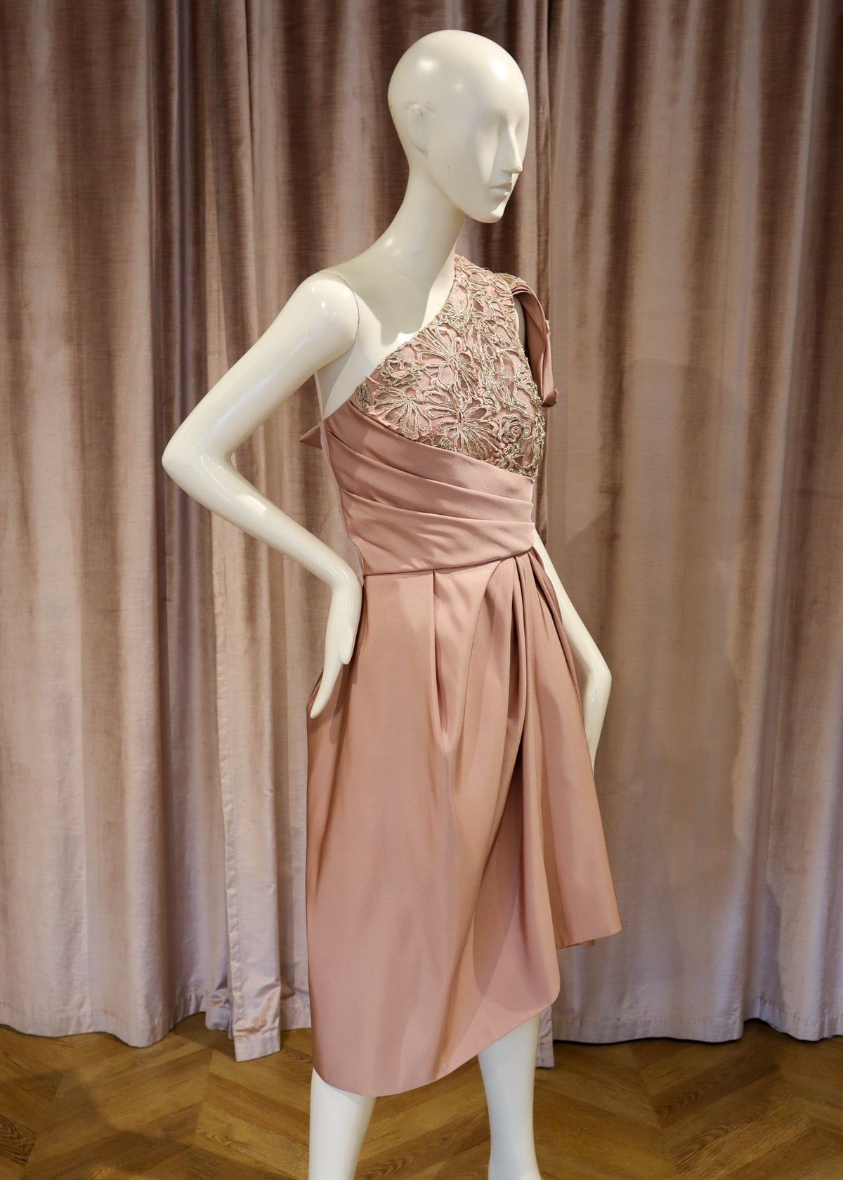 A one-shoulder dusty pink dress with a beaded bodice and a tied waist sash, displayed on a white mannequin against a mauve velvet curtain and wooden parquet floor.