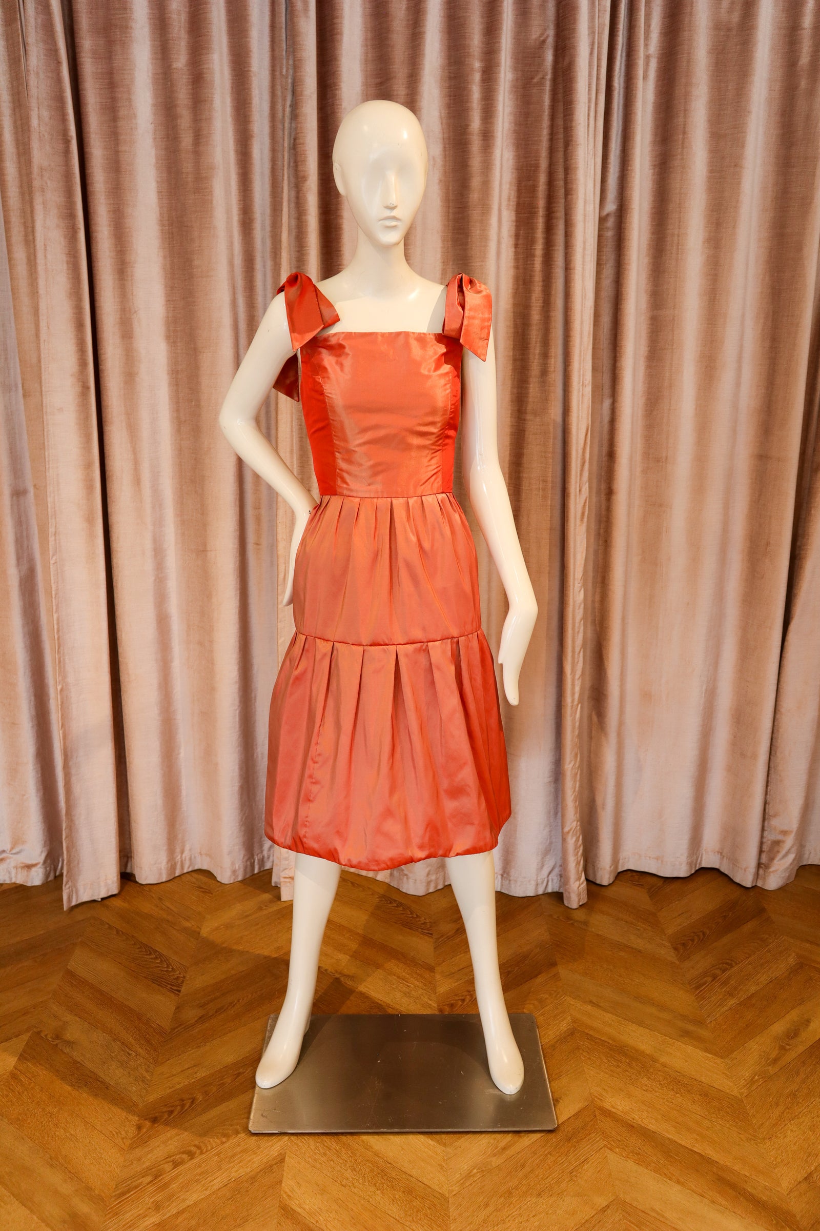 A bright coral-orange tiered dress displayed on a white mannequin, set against a backdrop of pinkish-beige curtains and a wooden herringbone floor.