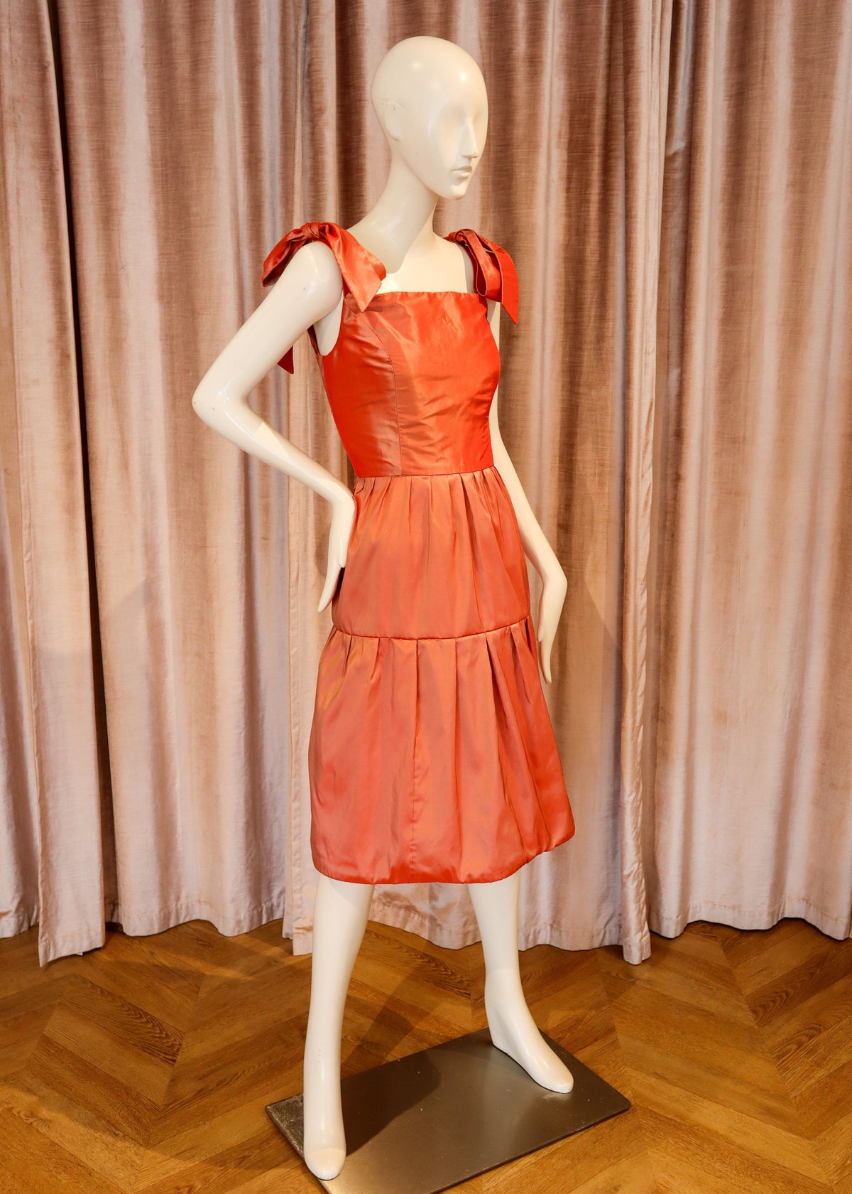 A bright coral-orange tiered dress displayed on a white mannequin, set against a backdrop of pinkish-beige curtains and a wooden herringbone floor.