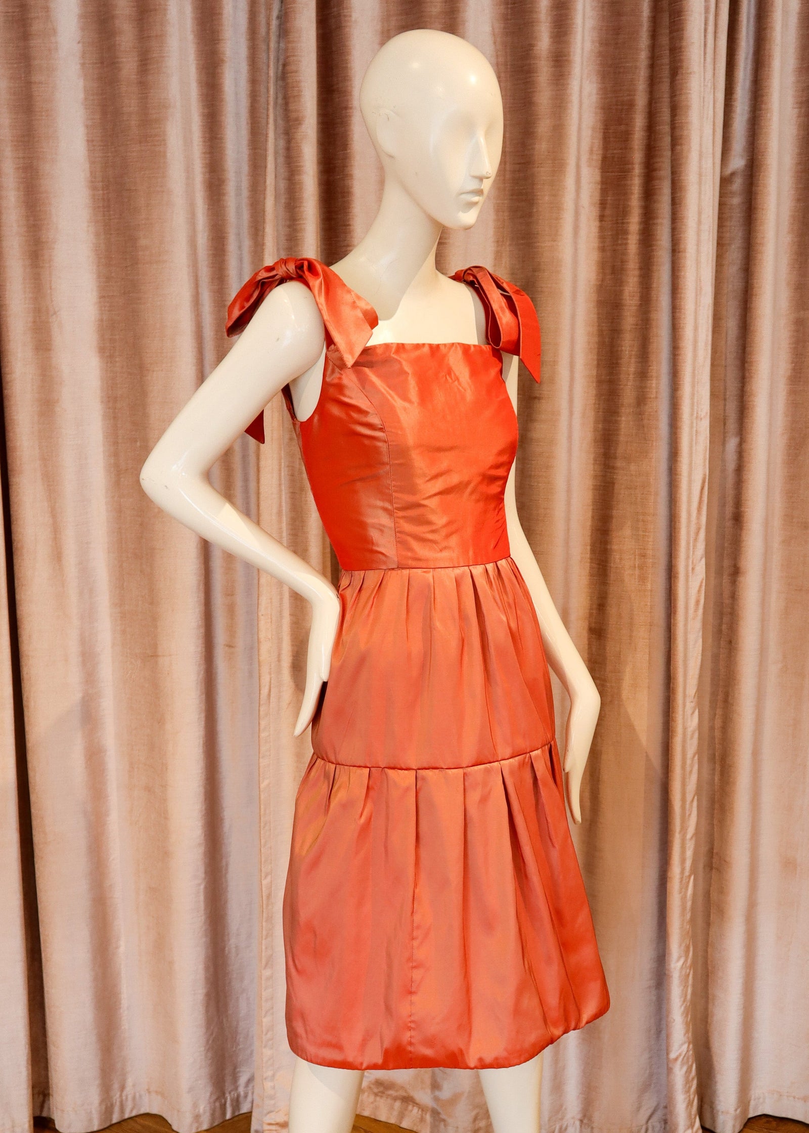 A bright coral-orange tiered dress displayed on a white mannequin, set against a backdrop of pinkish-beige curtains and a wooden herringbone floor.