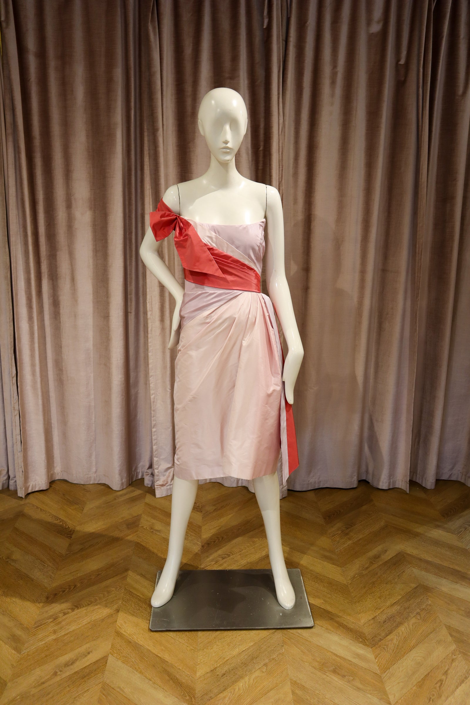 A white mannequin wearing a one-shoulder pink dress with a contrasting red bow and sash, standing on a black base against a brown curtain backdrop.