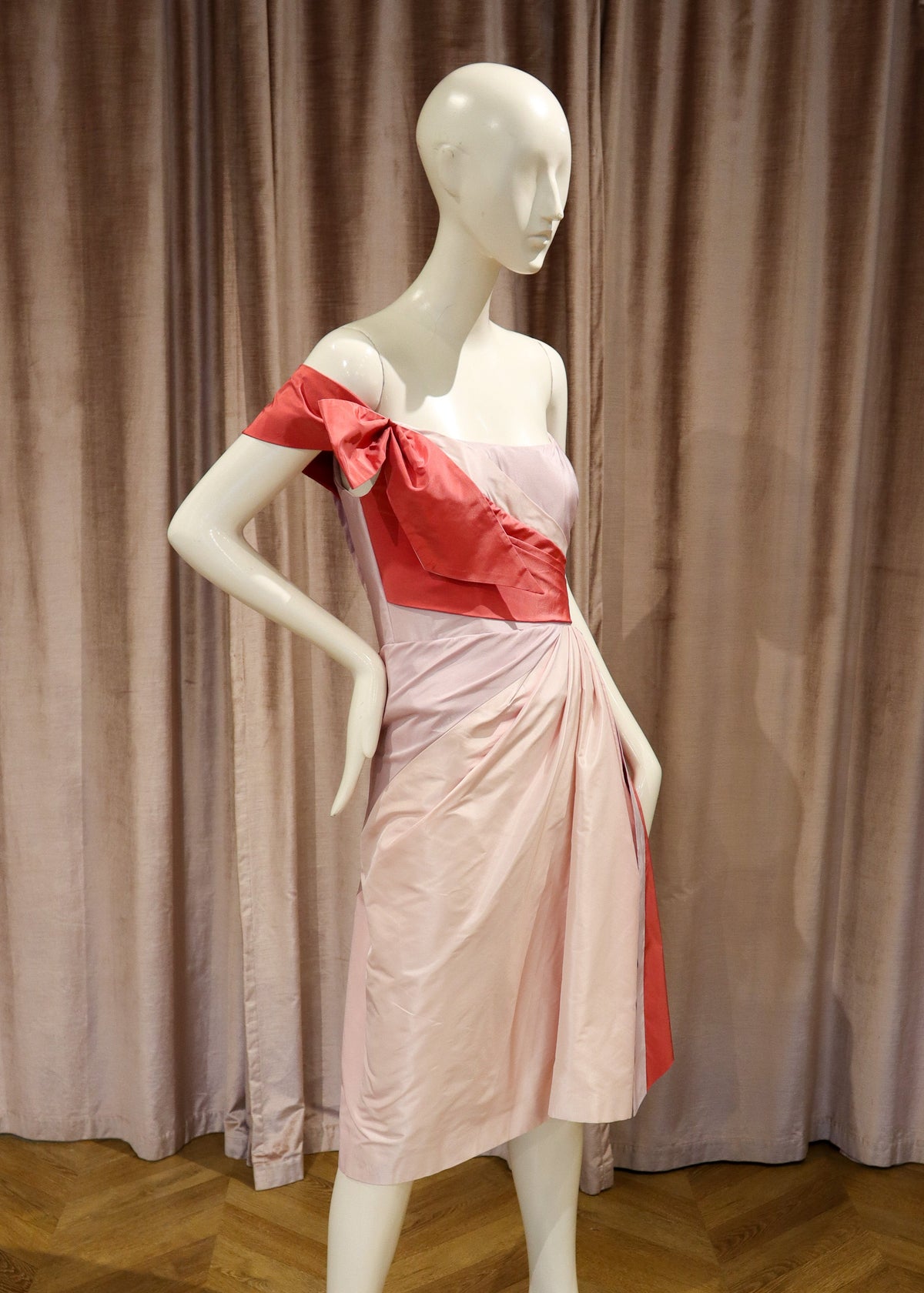 A white mannequin wearing a one-shoulder pink dress with a contrasting red bow and sash, standing on a black base against a brown curtain backdrop.