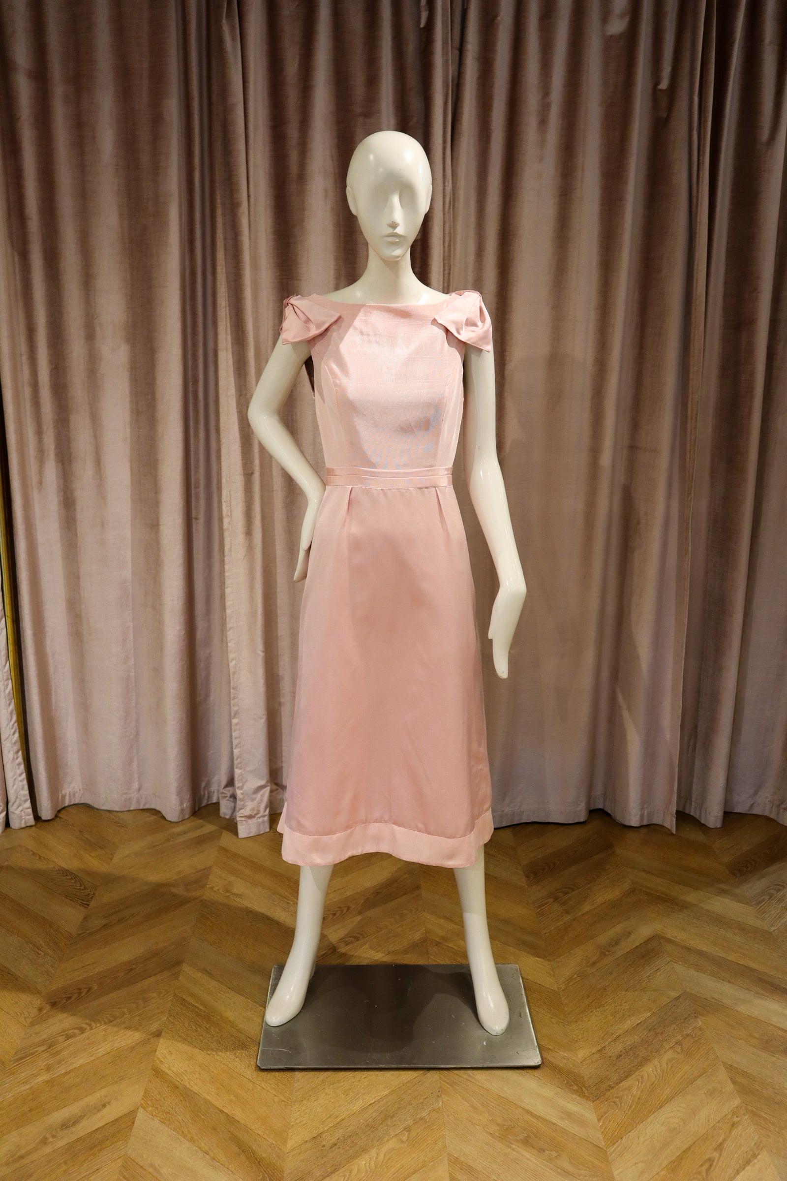 A light pink sleeveless midi dress displayed on a white mannequin, set against a backdrop of dusty rose velvet curtains and a wooden herringbone floor.