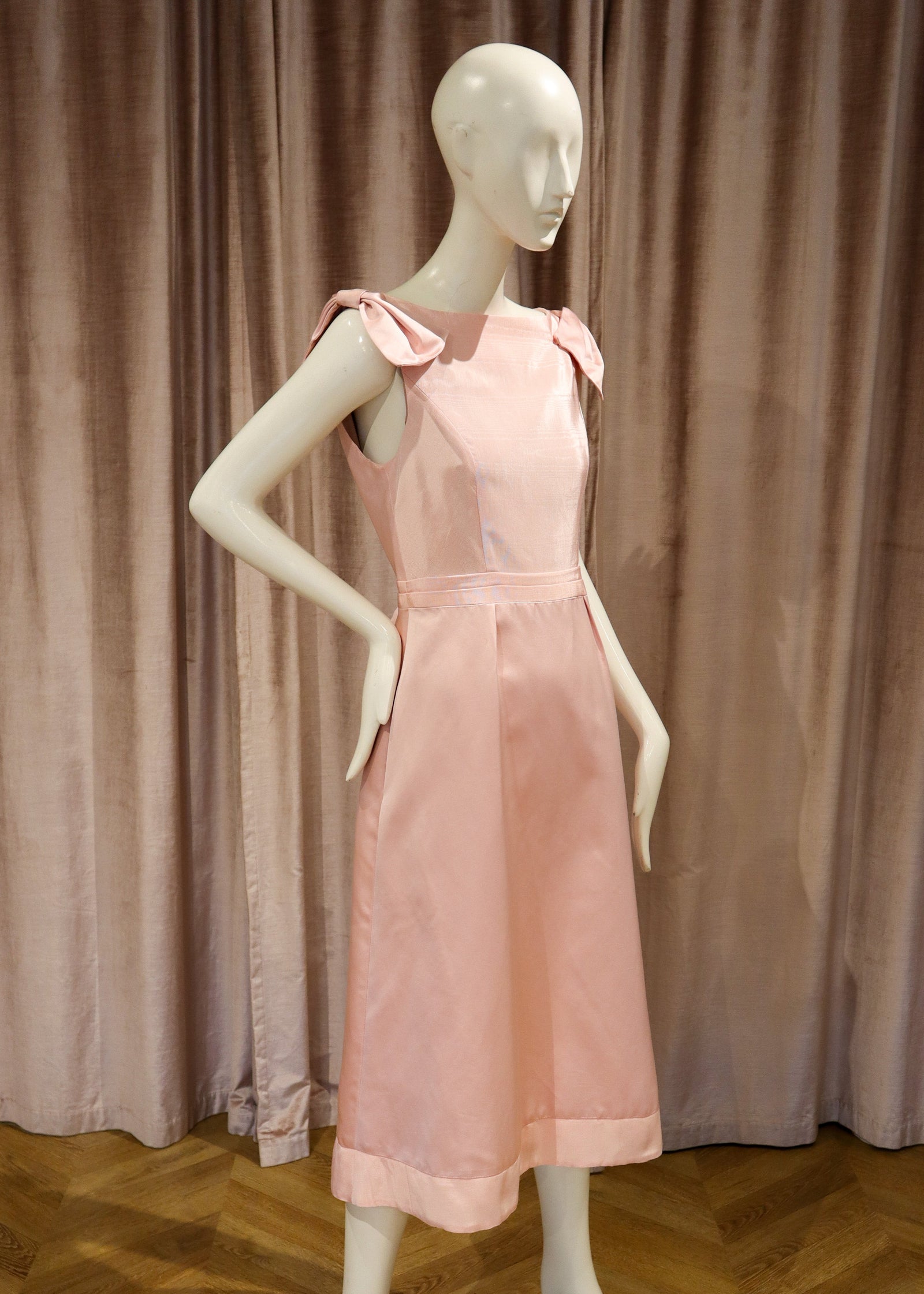 A light pink sleeveless midi dress displayed on a white mannequin, set against a backdrop of dusty rose velvet curtains and a wooden herringbone floor.
