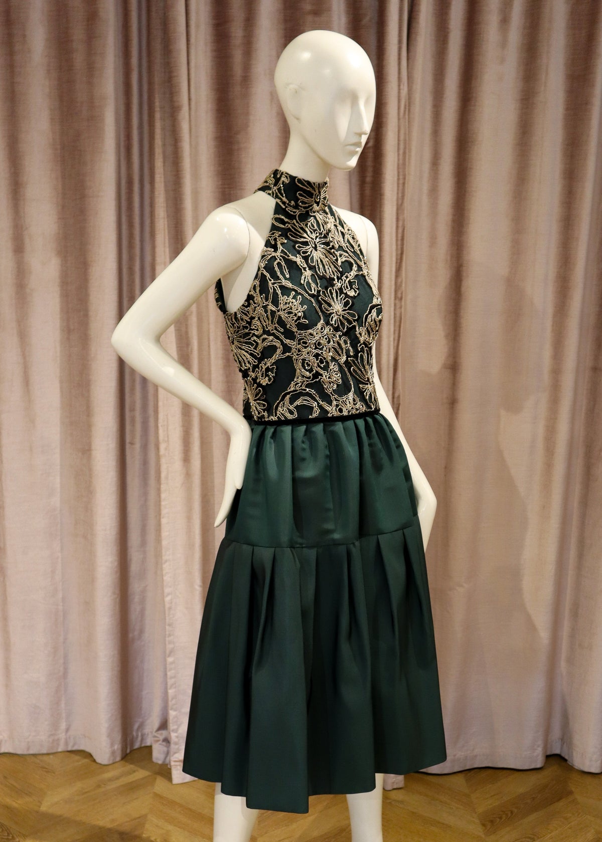 A sleeveless high-neck dress displayed on a white mannequin, featuring a black and gold floral embroidered bodice and a dark green tiered skirt, set against a dusty pink curtain backdrop.