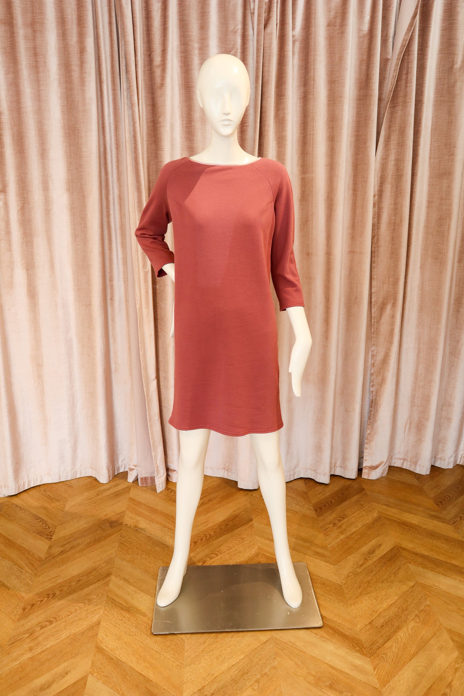 A terracotta-colored three-quarter sleeve dress displayed on a white mannequin, set against a backdrop of pinkish-beige curtains and a wooden herringbone floor.