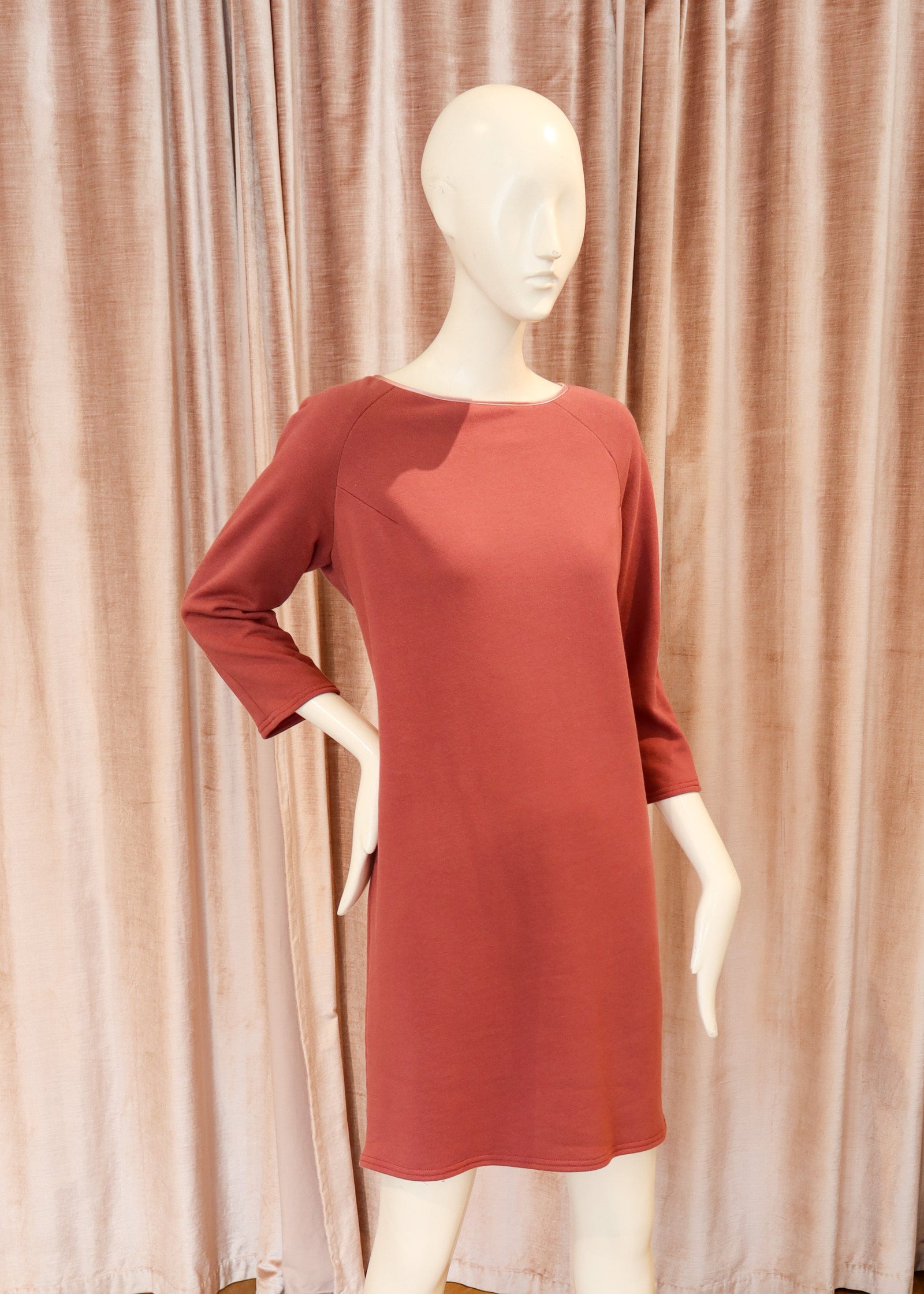 A terracotta-colored three-quarter sleeve dress displayed on a white mannequin, set against a backdrop of pinkish-beige curtains and a wooden herringbone floor.