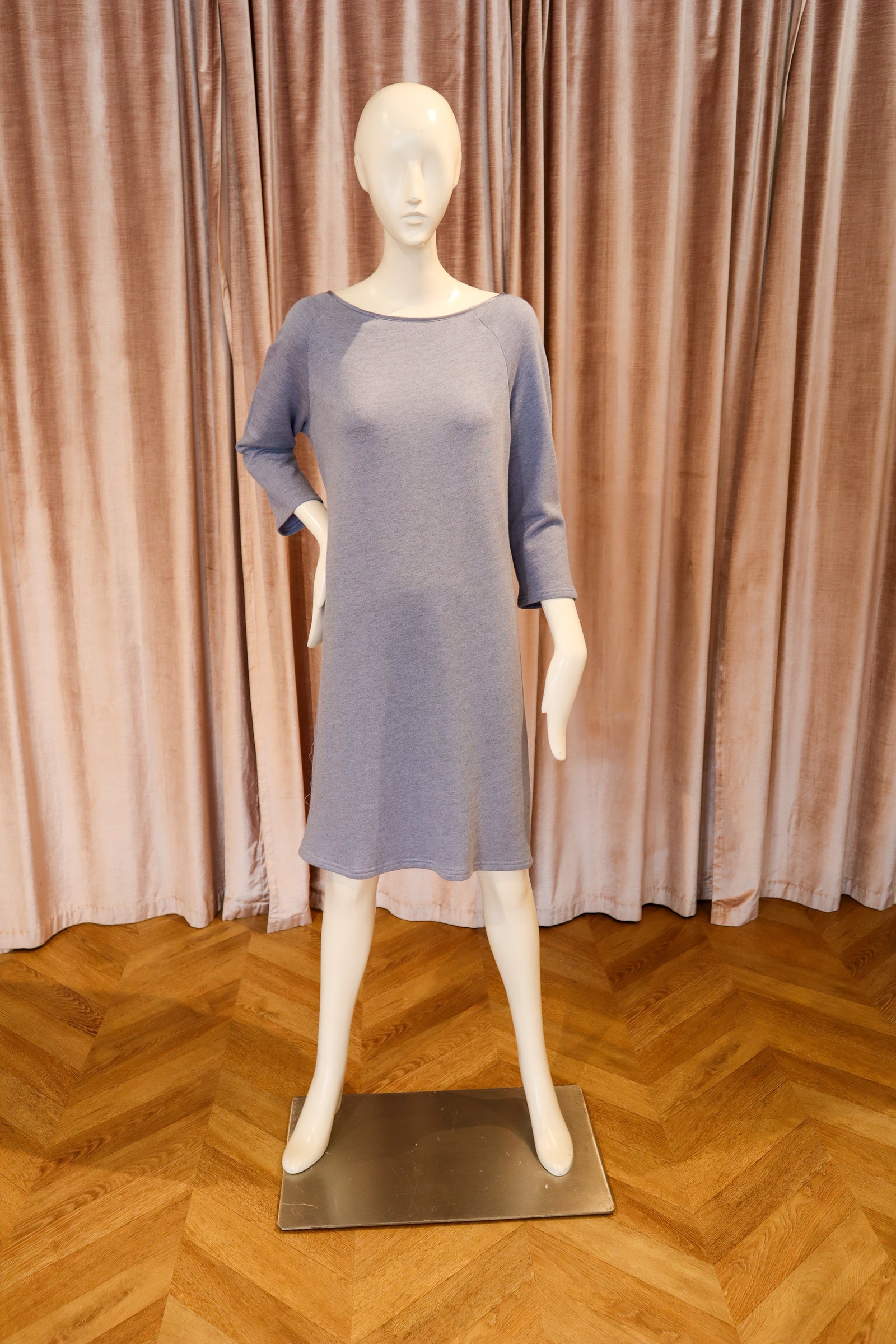 A three-quarter sleeve knit dress in a muted dusty lavender-blue color, displayed on a white mannequin with a metal base against a backdrop of pinkish-brown velvet curtains and a wooden herringbone floor.