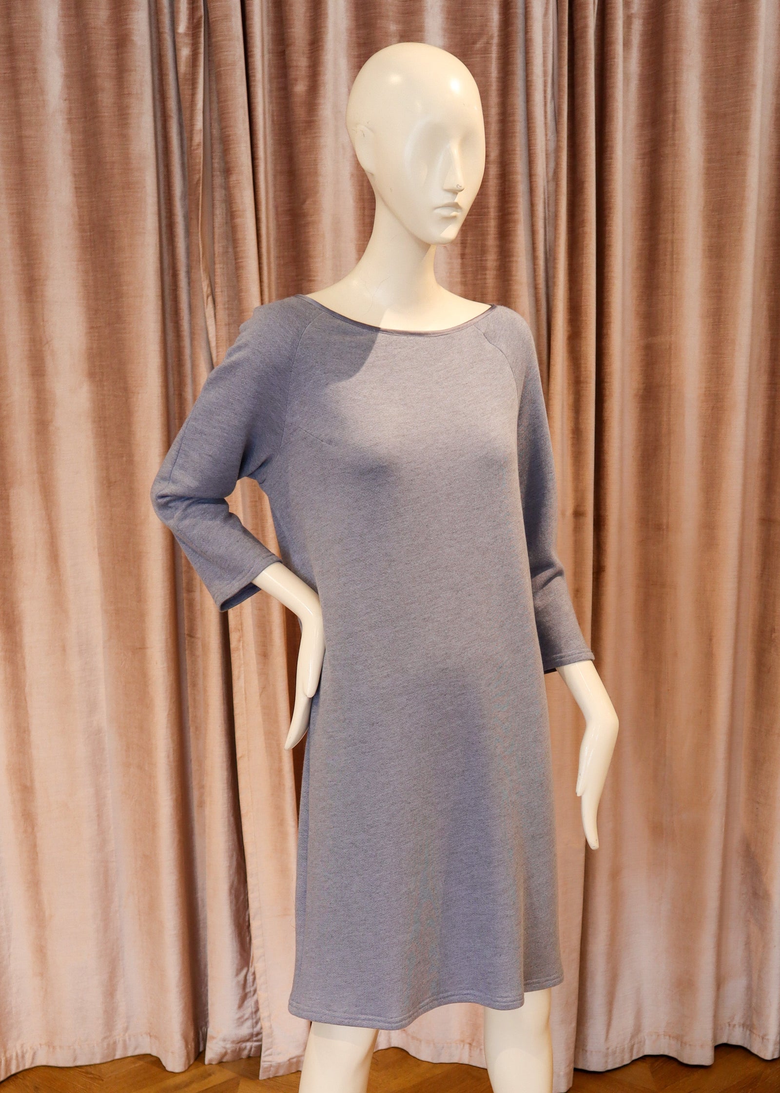 A three-quarter sleeve knit dress in a muted dusty lavender-blue color, displayed on a white mannequin with a metal base against a backdrop of pinkish-brown velvet curtains and a wooden herringbone floor.