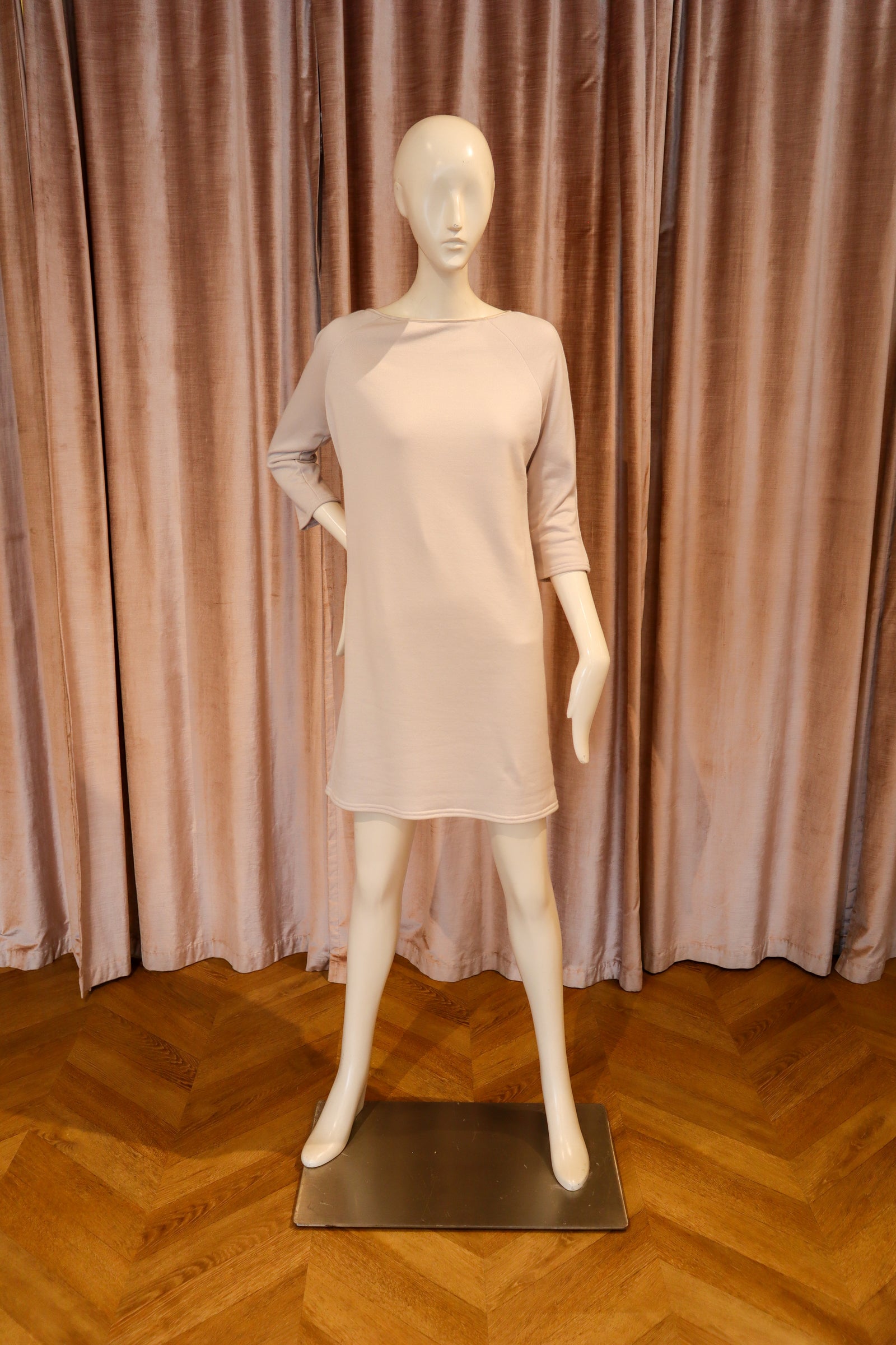 A pale blush pink long-sleeve mini dress displayed on a white female mannequin, set against a backdrop of dusty rose curtains and a wooden parquet floor.