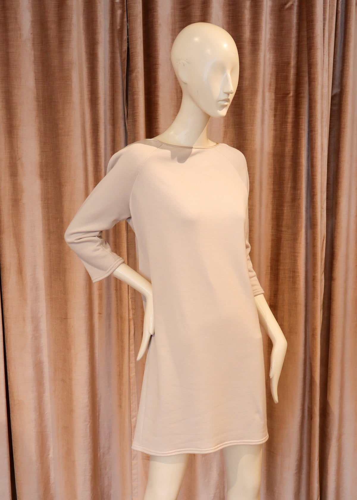A pale blush pink long-sleeve mini dress displayed on a white female mannequin, set against a backdrop of dusty rose curtains and a wooden parquet floor.