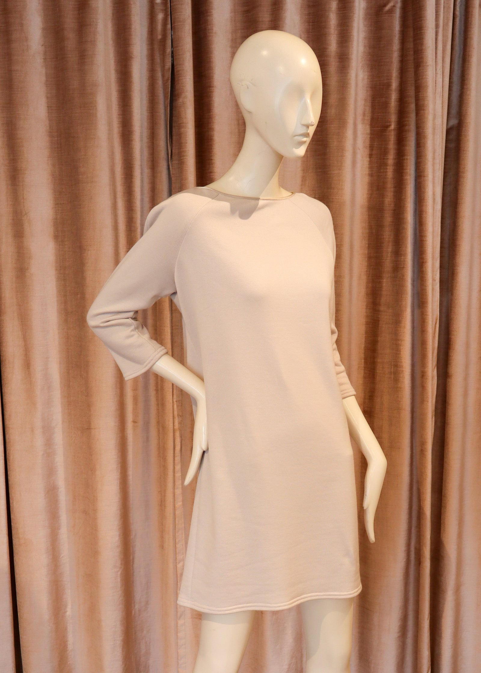 A pale blush pink long-sleeve mini dress displayed on a white female mannequin, set against a backdrop of dusty rose curtains and a wooden parquet floor.