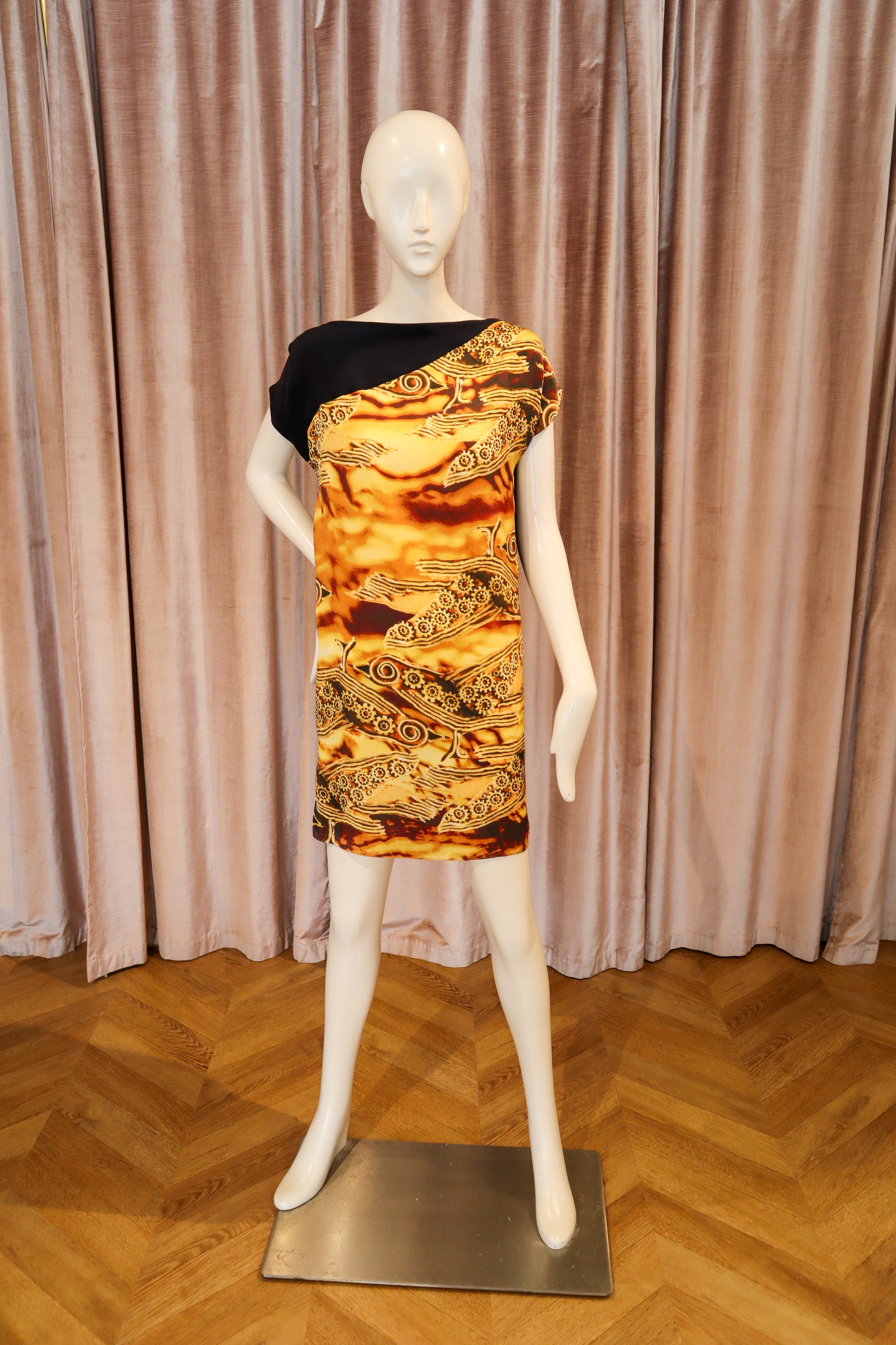 A one-shoulder mini dress displayed on a white mannequin, featuring a black shoulder panel and a vibrant gold, orange, and brown patterned body, set against a pink curtain and wooden floor background.
