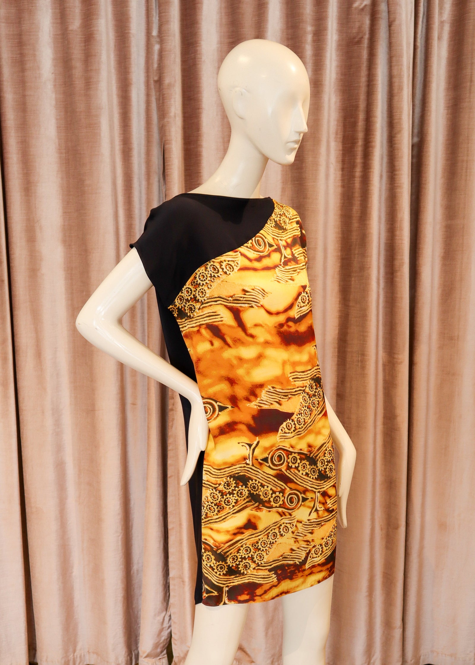 A one-shoulder mini dress displayed on a white mannequin, featuring a black shoulder panel and a vibrant gold, orange, and brown patterned body, set against a pink curtain and wooden floor background.